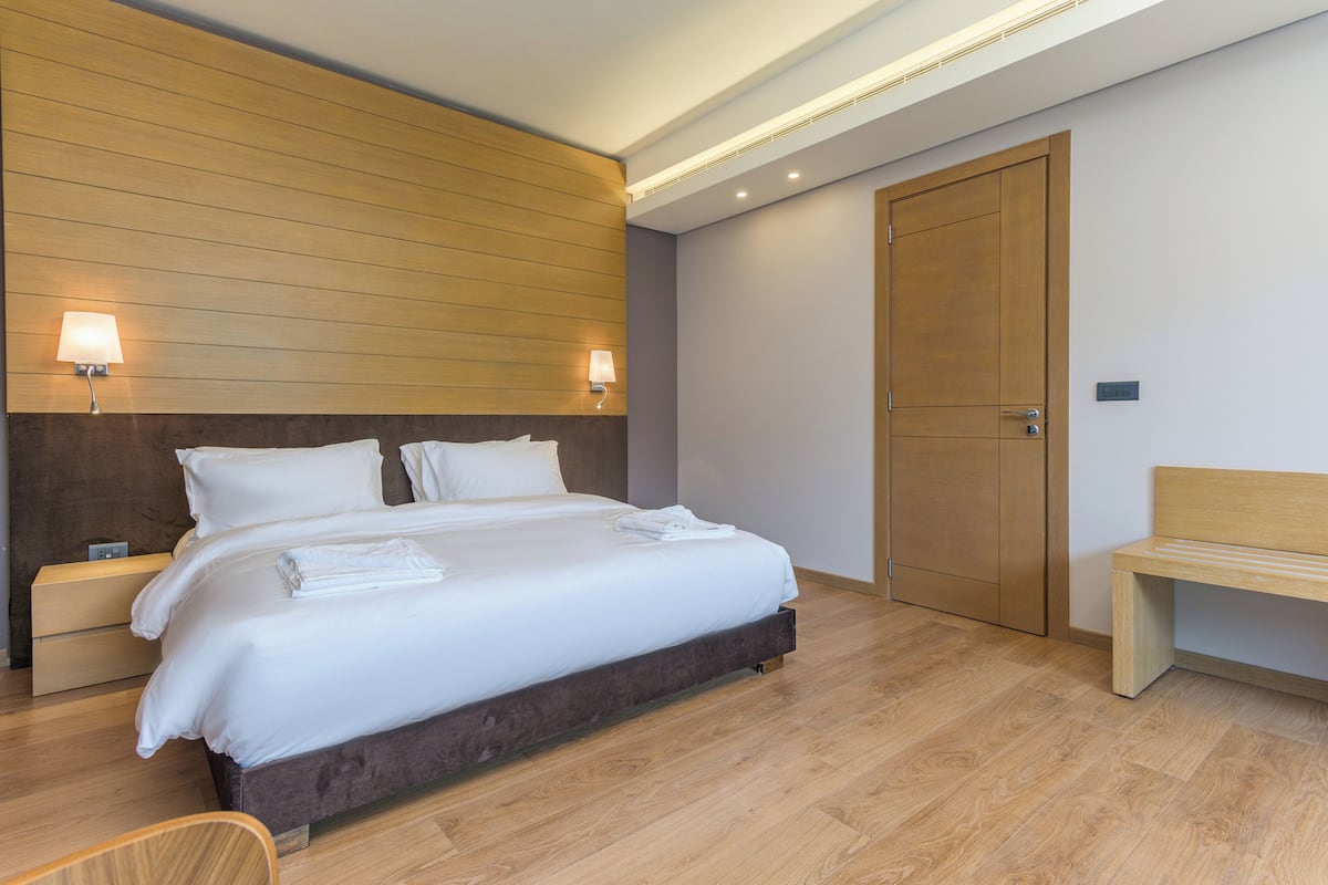 A modern hotel room with a large bed, wooden headboard, two bedside lamps, a wooden bench, and a wardrobe.