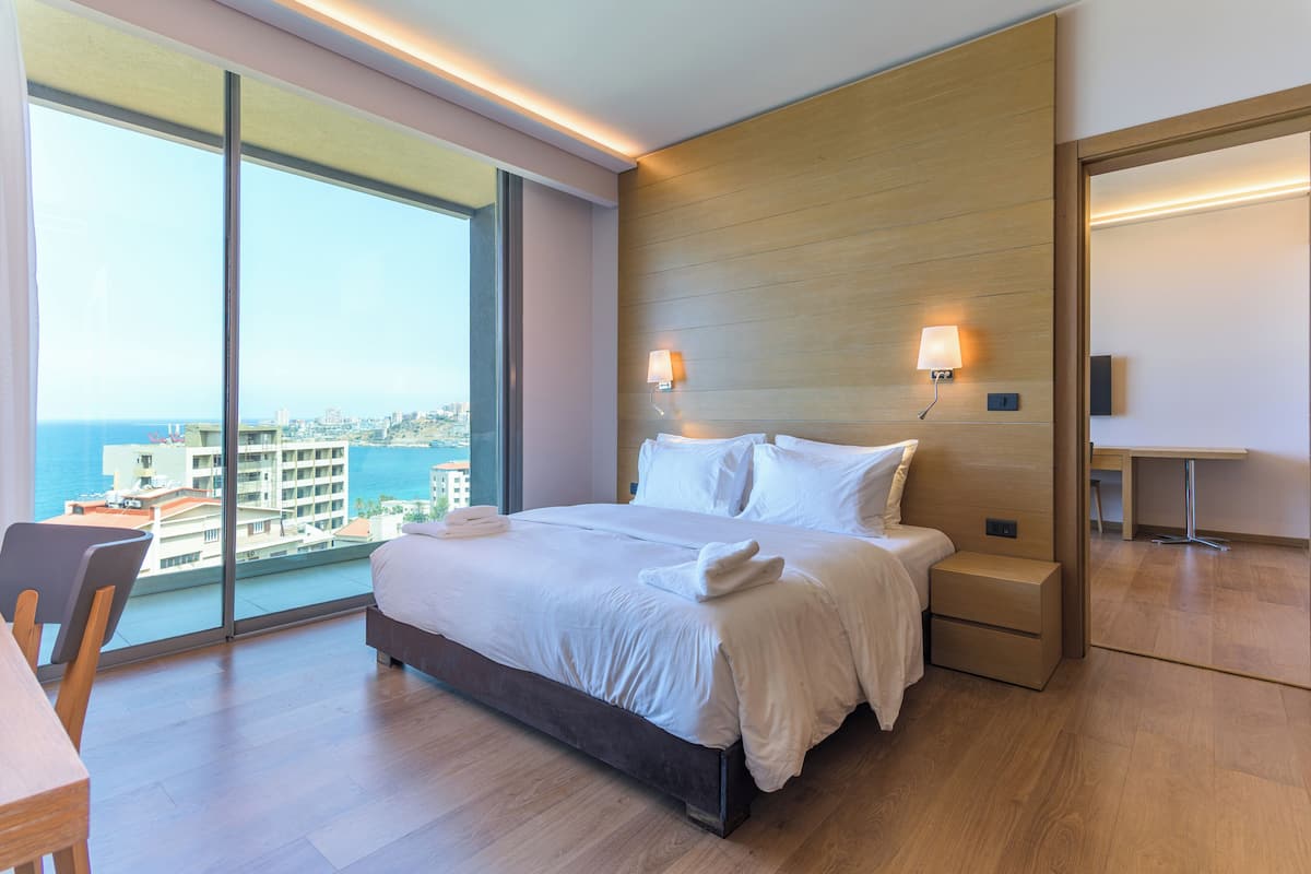 A modern hotel room with a large bed, bedside tables, a desk, and a view of the city and sea.