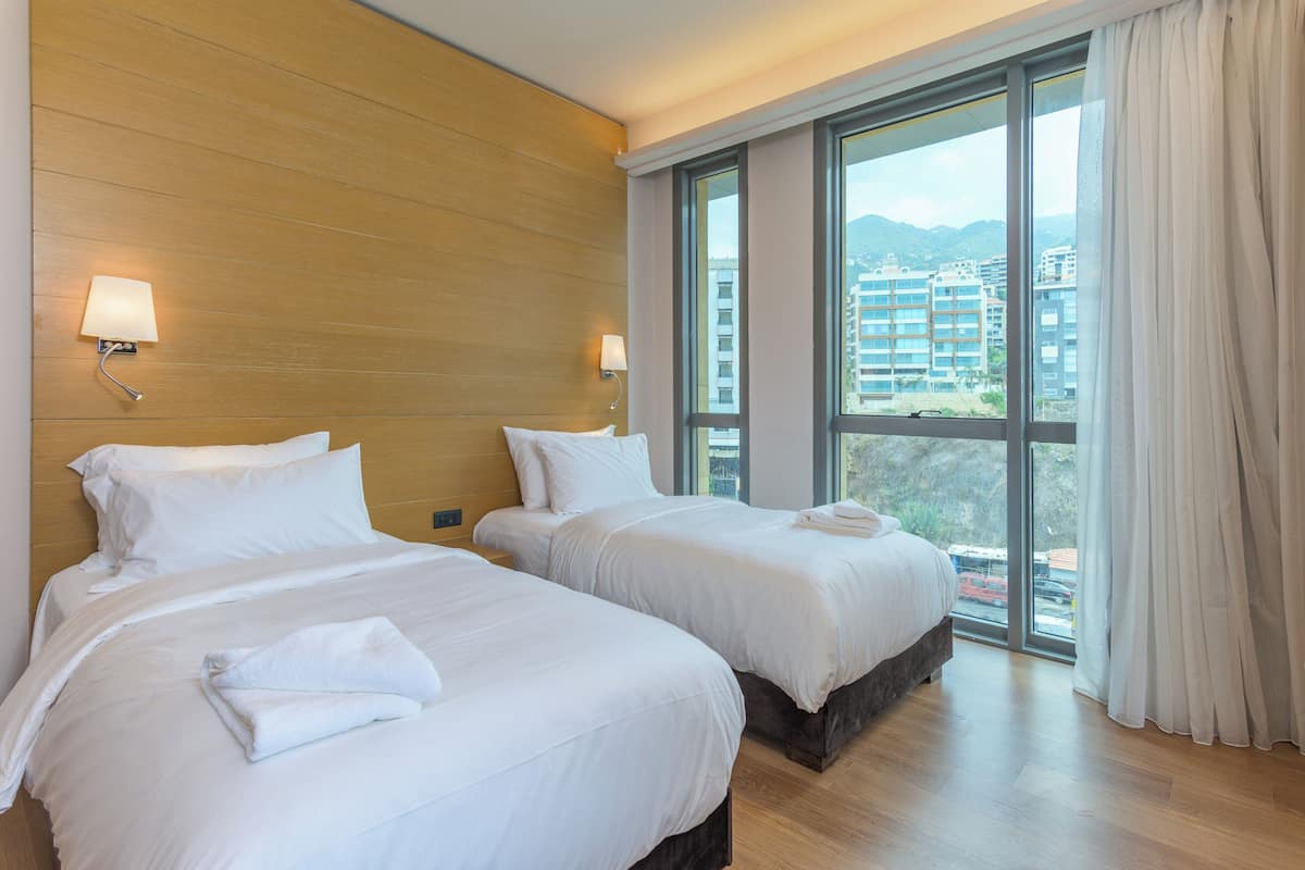 A hotel room with two beds, wooden headboard, large windows, and a view of buildings.