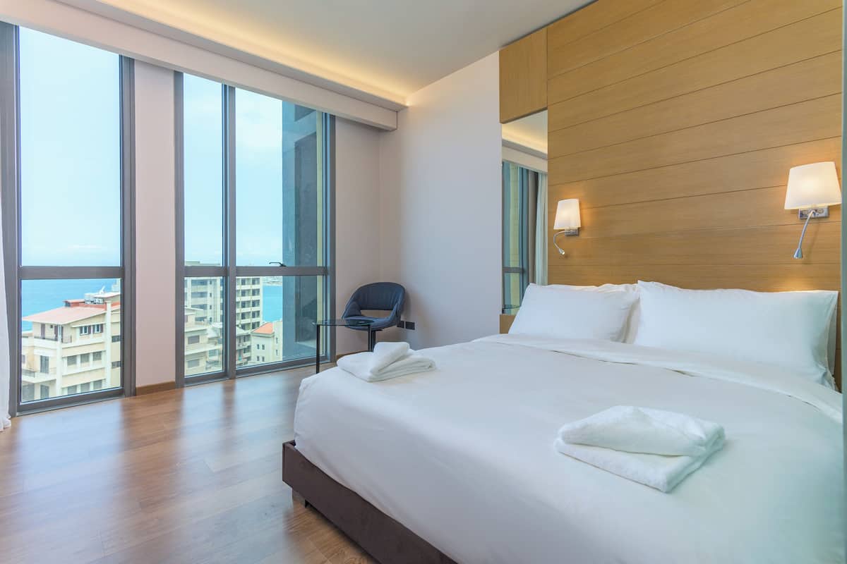 A hotel room with a large bed, a chair, and a view of the city through floor-to-ceiling windows.