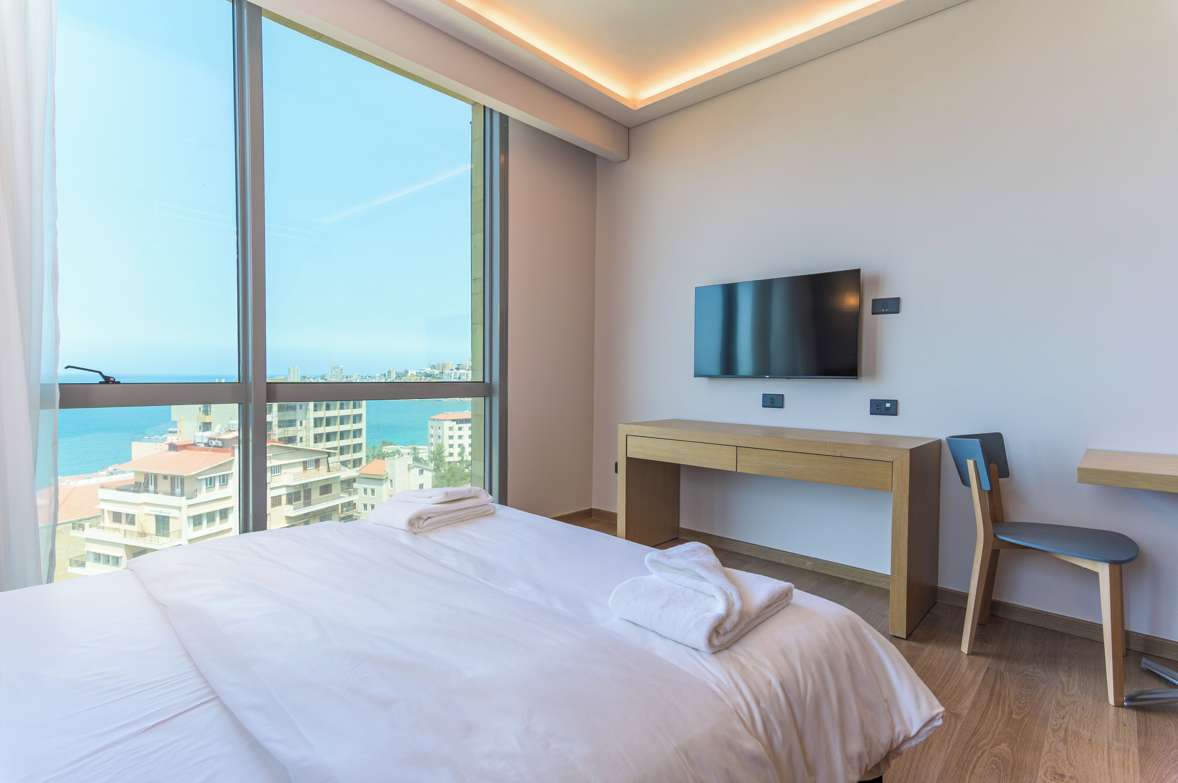 Ein Hotelzimmer mit großem Fenster, einem Bett, einem Schreibtisch, einem Sessel und einem Flachbildfernseher.