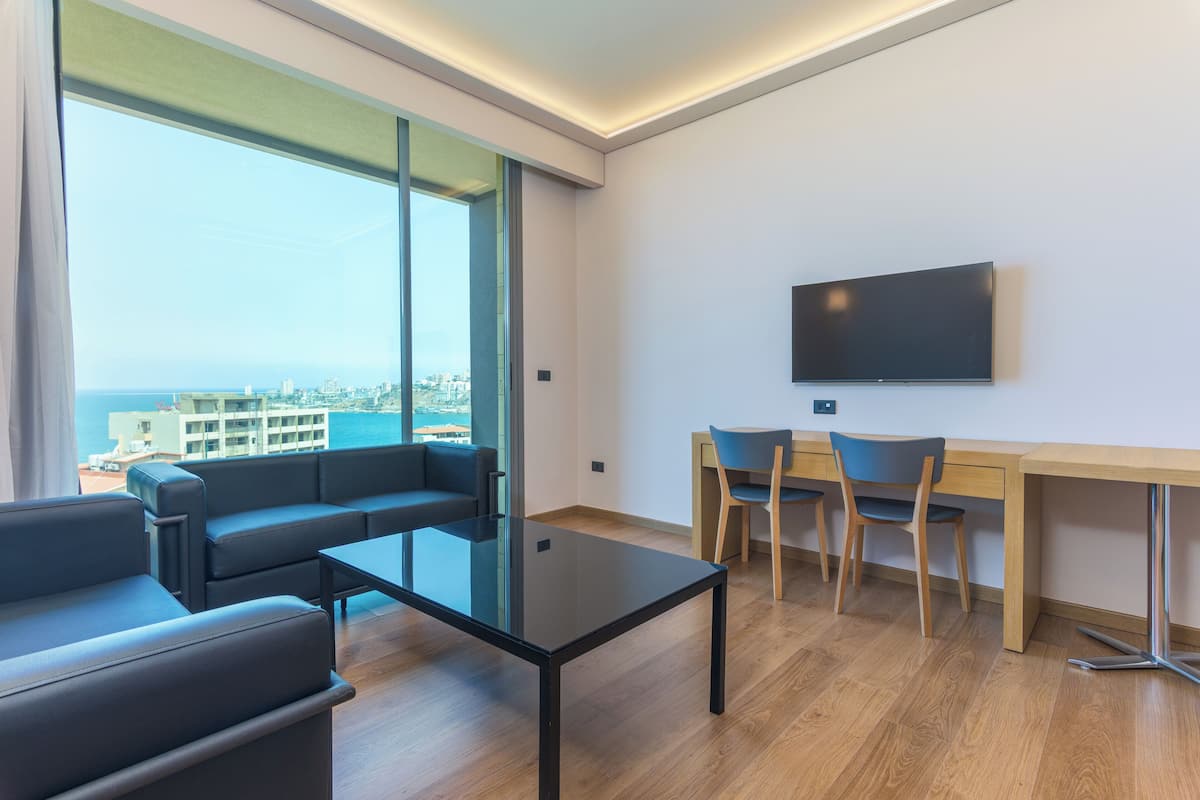 A modern living room with a glass wall, a sofa, a coffee table, a TV, and a desk with chairs.