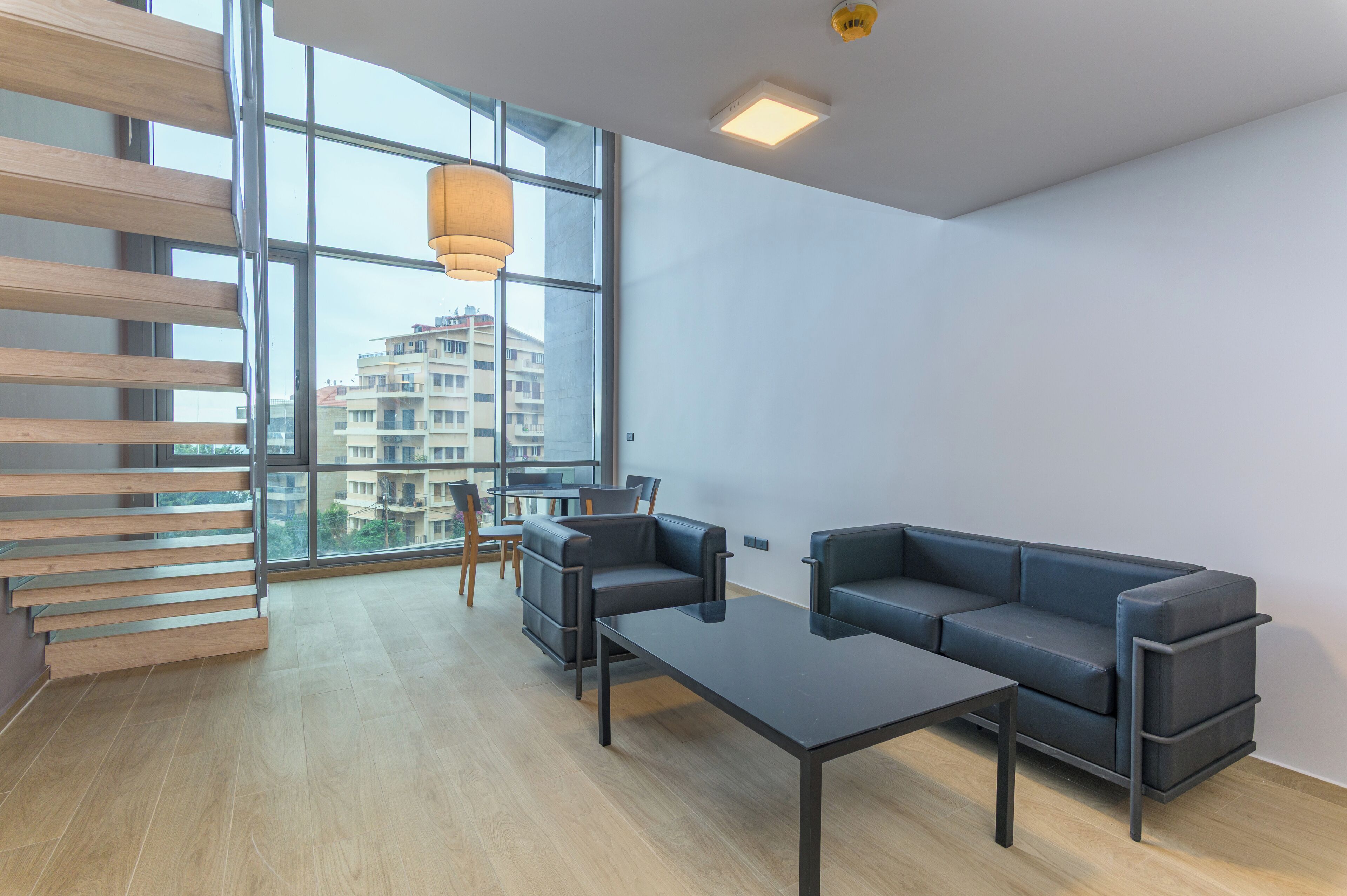 A modern living room with a sofa, coffee table, and a dining area with chairs and a table. Large windows provide a view of buildings outside.