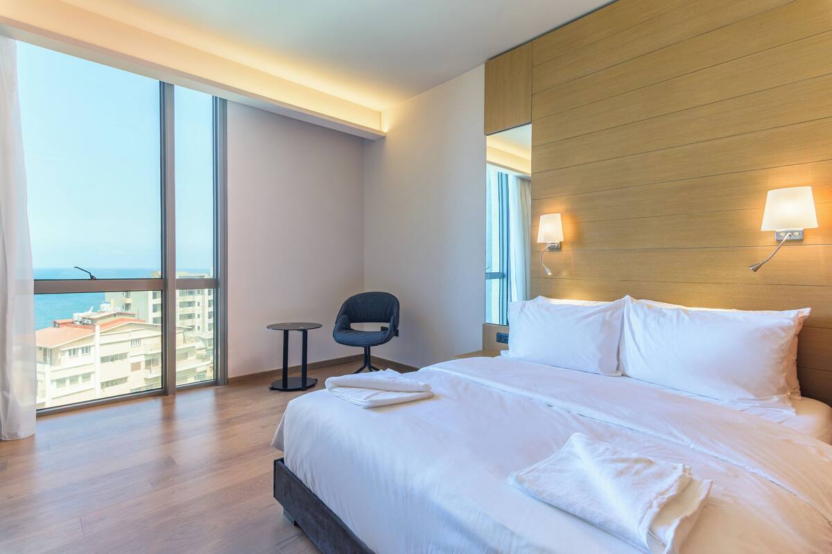 A hotel room with a large bed, two bedside lamps, a chair, and a view of the city through large windows.