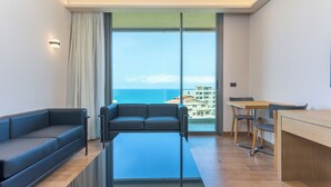 A modern living room with a sofa, a glass coffee table, a dining table with chairs, and a view of the ocean through large windows. - Jounieh Living - Residences by Local Host (Jounieh)