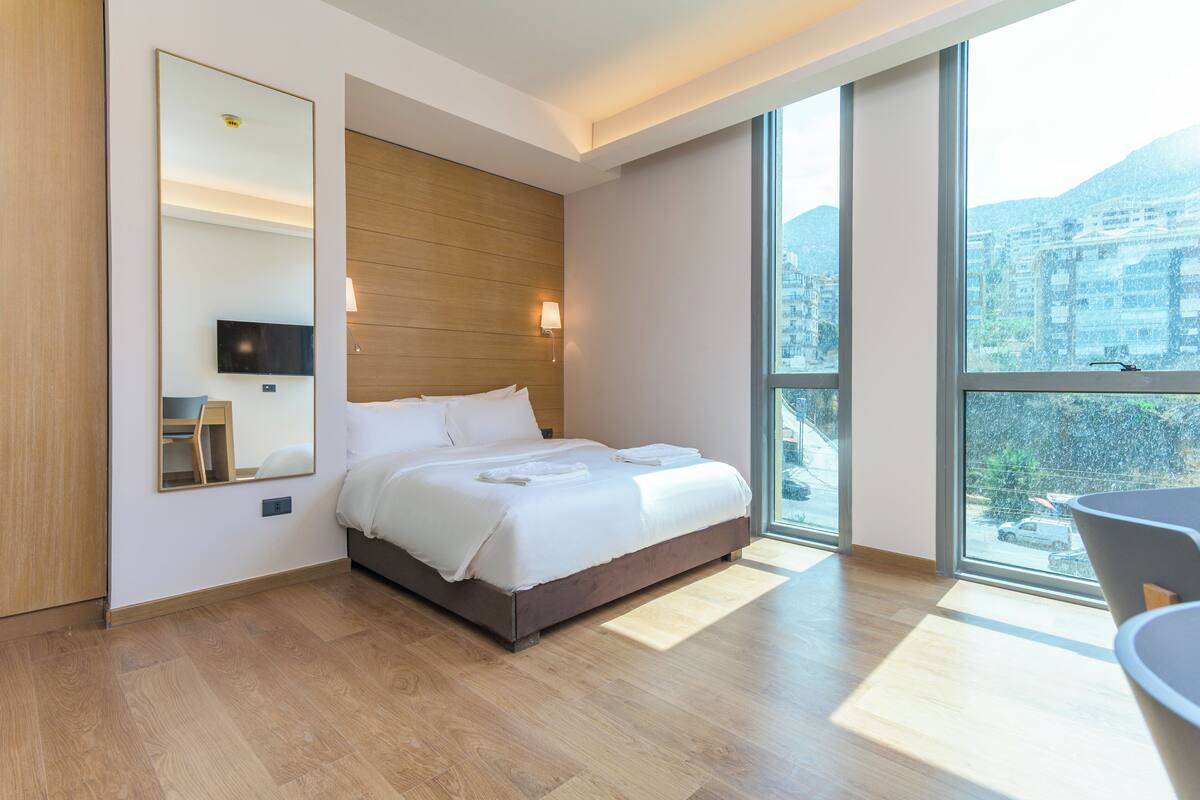 A modern hotel room with a large bed, a TV, a wooden headboard, a large window with a city view, and a small table with chairs.