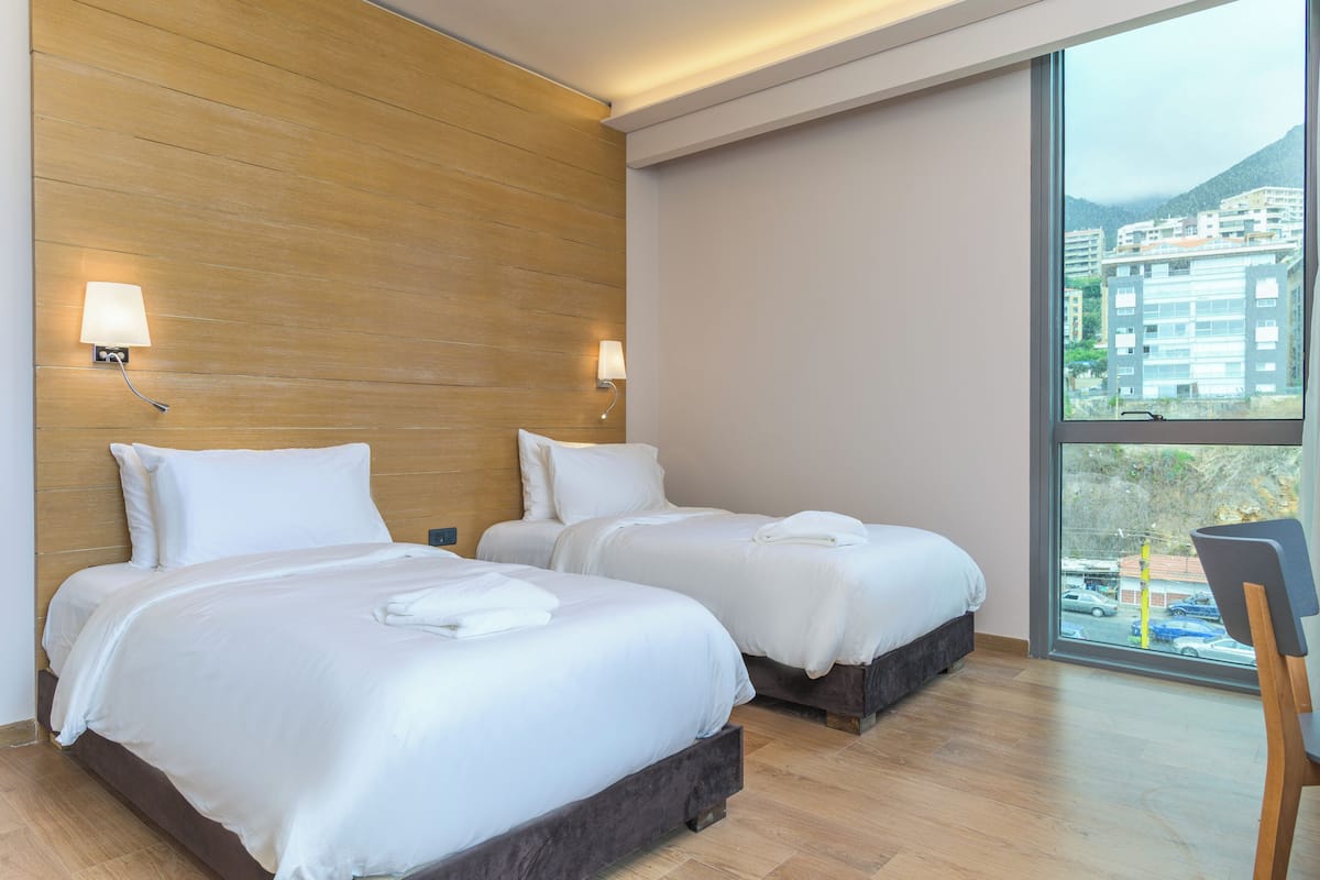 A hotel room with two beds, wooden headboard, bedside lamps, and a large window with a view of buildings and mountains.