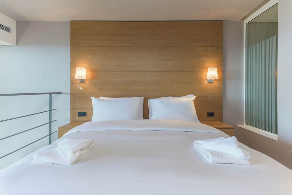 A hotel room with a wooden headboard, two bedside lamps, a bed with white linens, and a window with blinds.