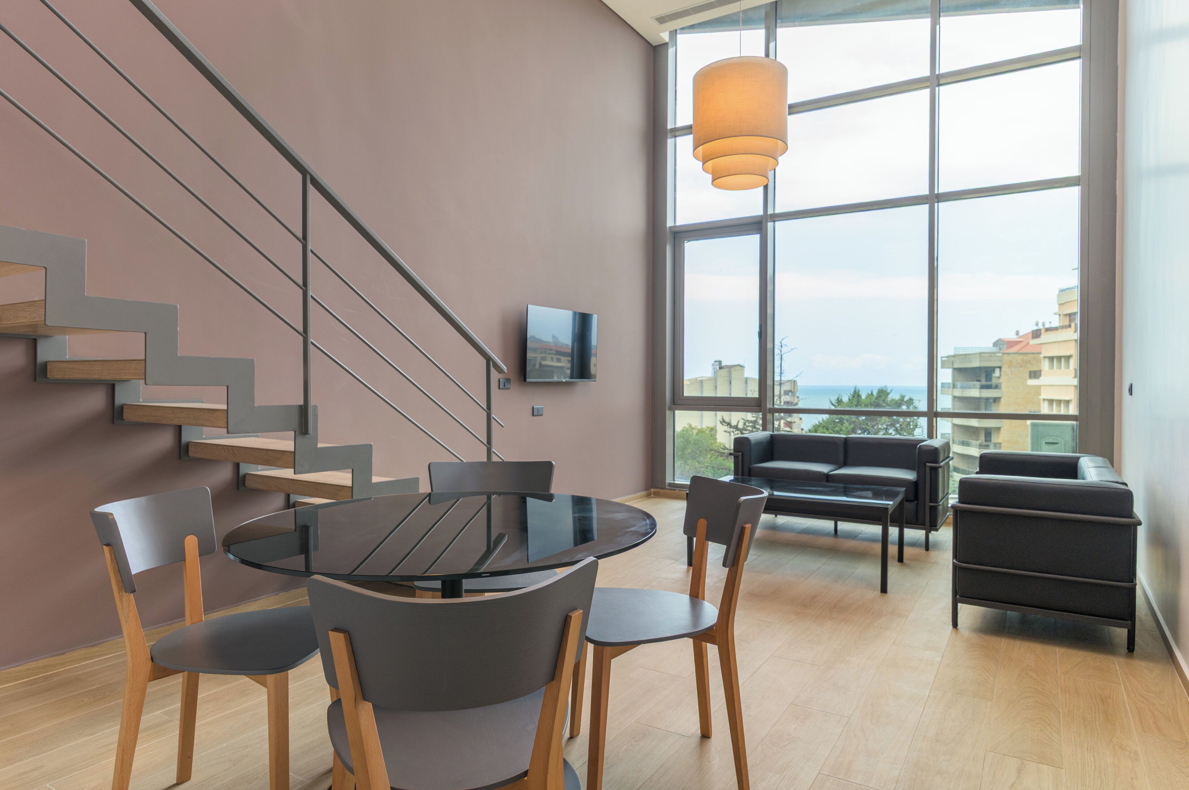 A modern interior with a staircase, a round table with chairs, a sofa, and a large window offering a view.