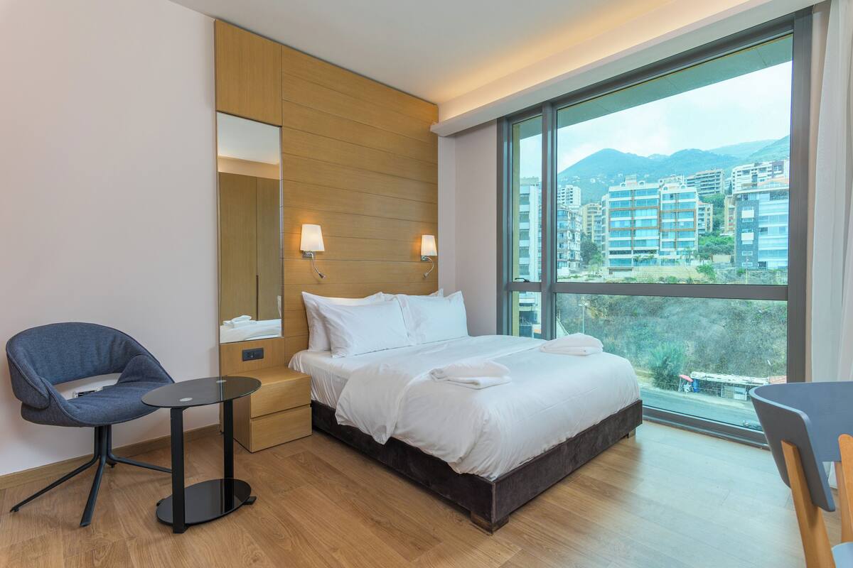 A hotel room with a large bed, a small table, a chair, and a large window offering a city view.
