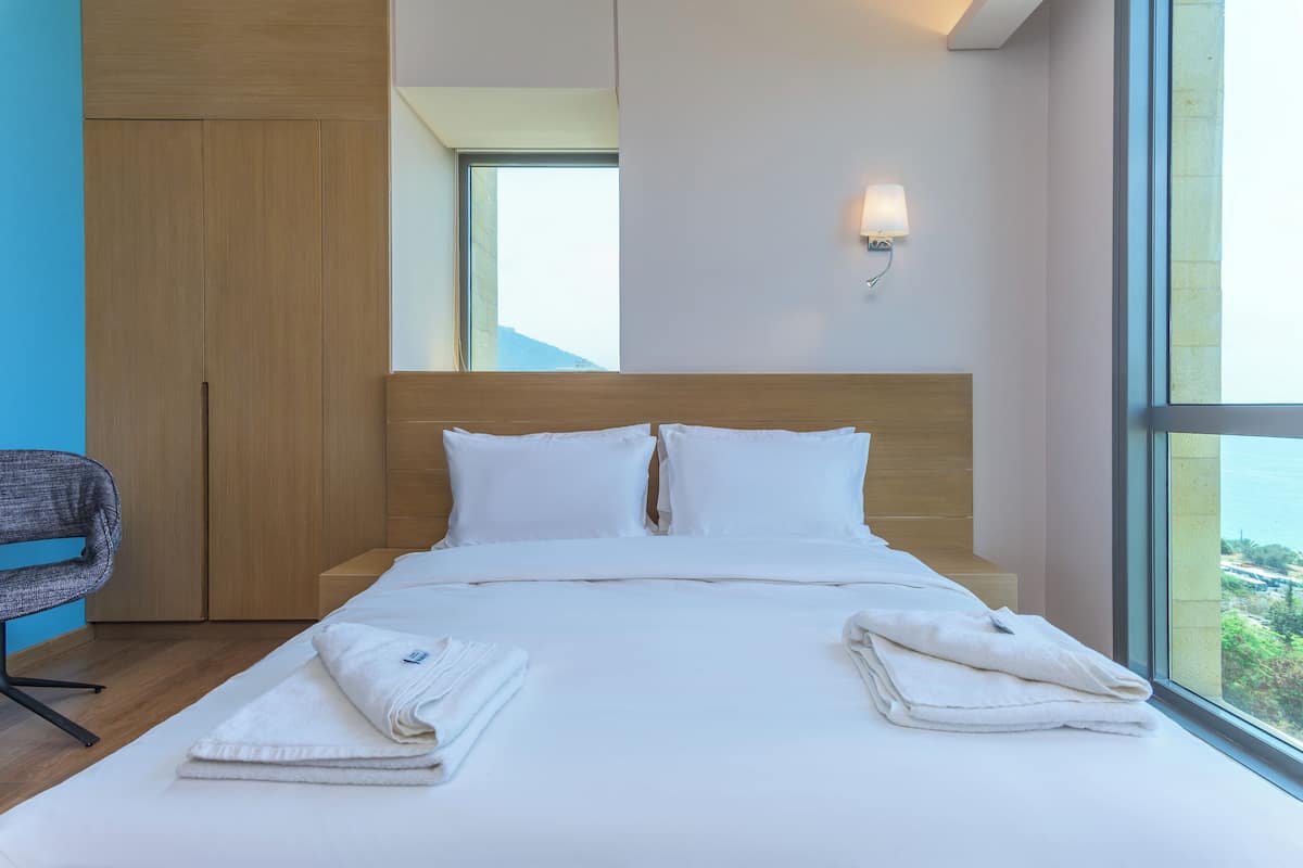 A modern hotel room with a large bed, a wooden wardrobe, a desk, and a view of the city through large windows.