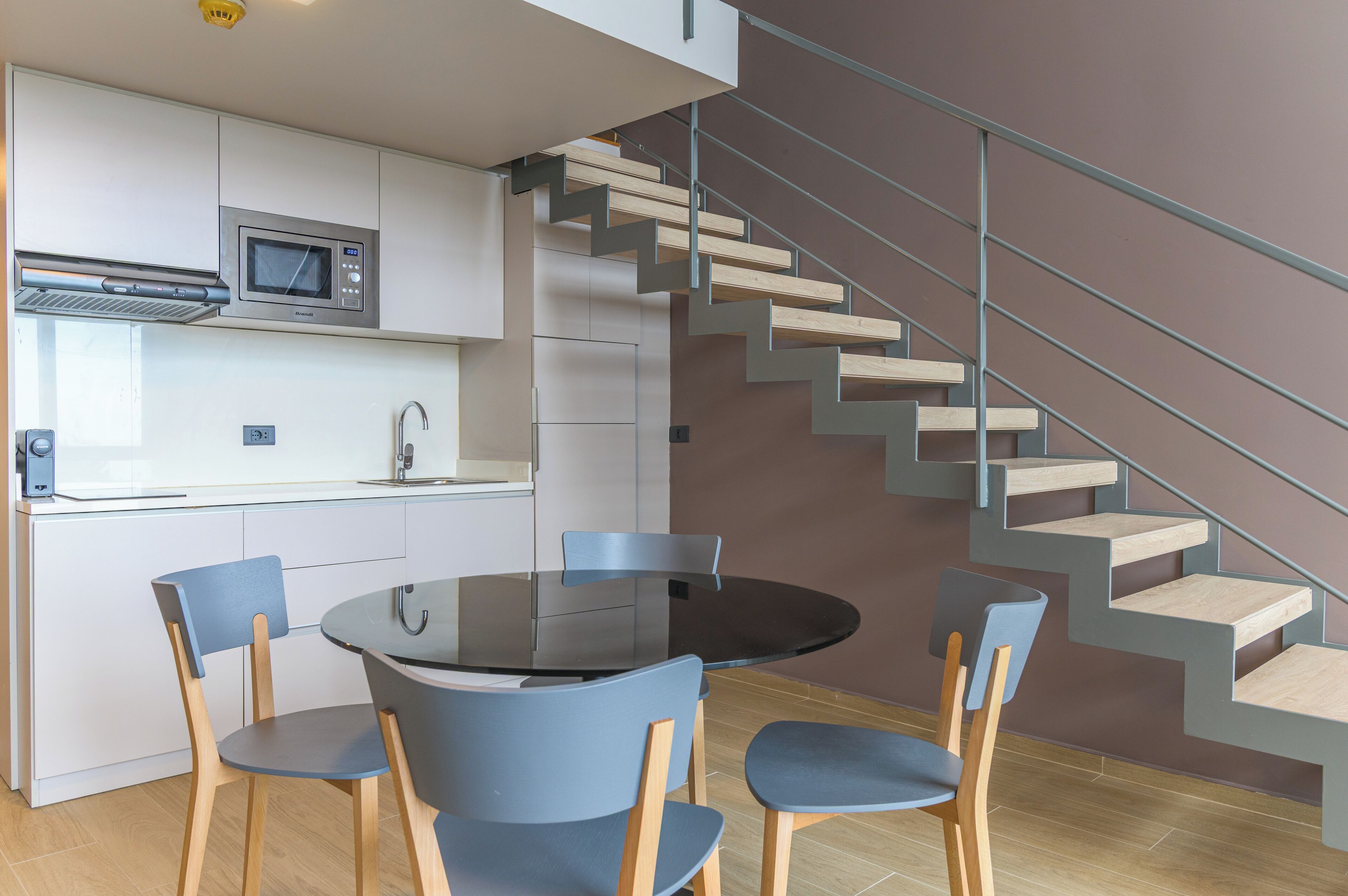 A modern kitchen with a built-in microwave, a dining area with a round table and four chairs, and a staircase with wooden steps and metal railings.