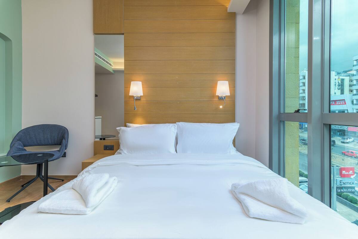 A hotel room with a bed, two wall-mounted lamps, a chair, and a view of the city through large windows.