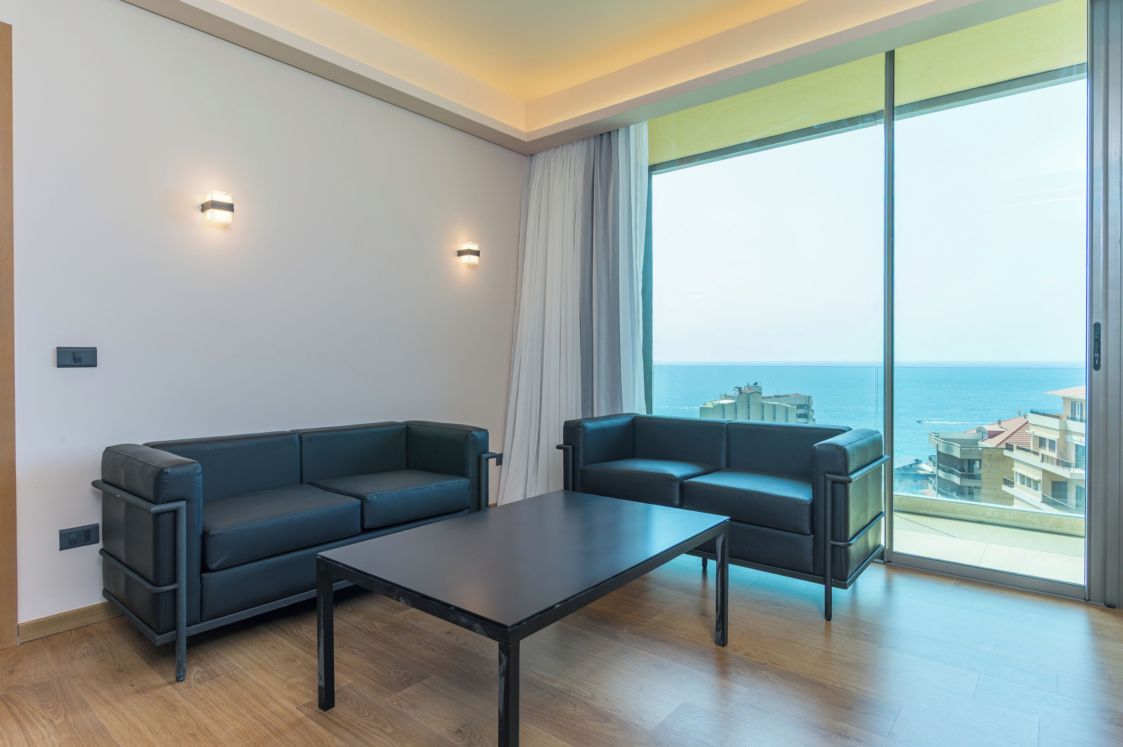 A modern living room with a sofa set, a coffee table, and a large window offering a view of the sea and buildings.