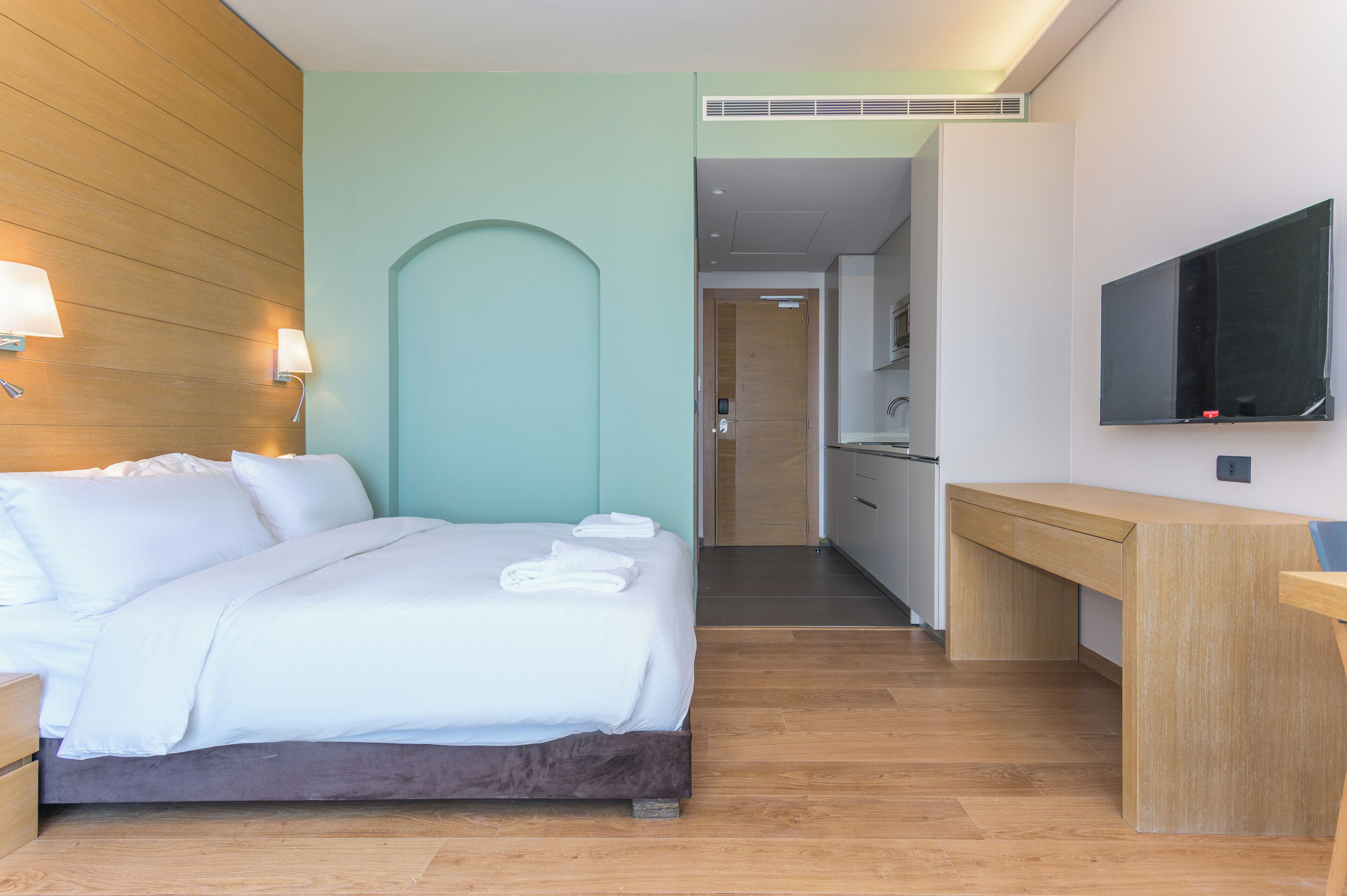 A modern hotel room with a bed, bedside lamps, a TV, a desk, and a bathroom visible through an archway.