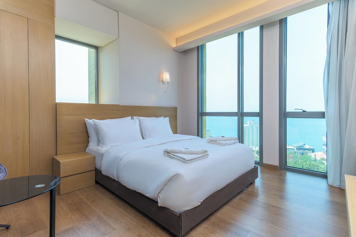 A modern hotel room with a large bed, a nightstand, a small table, and a view of the city through large windows.