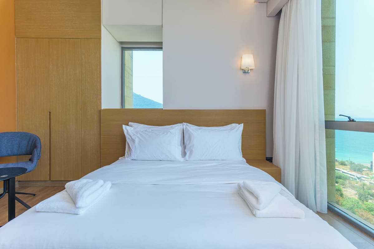 A hotel room with a bed, a chair, a window with a view, and a wall-mounted light.