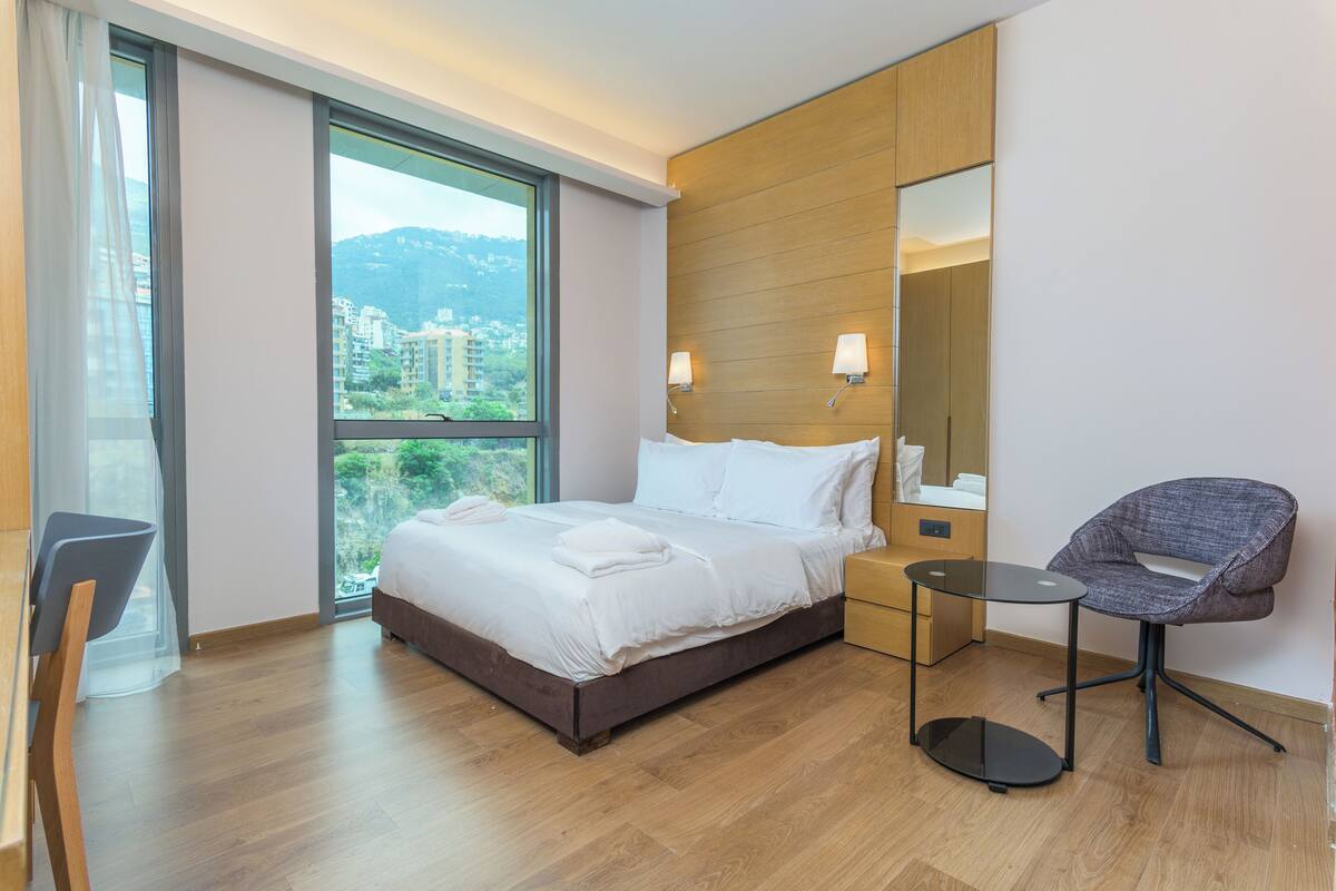 A modern hotel room with a large bed, a small table, a chair, and a large window offering a city view.