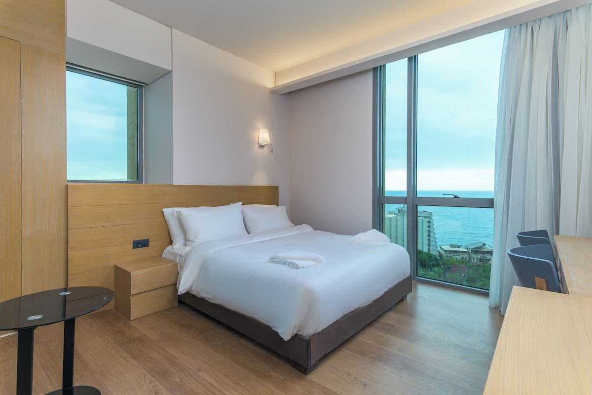 A modern hotel room with a large bed, a nightstand, a small table, and a view of the ocean.