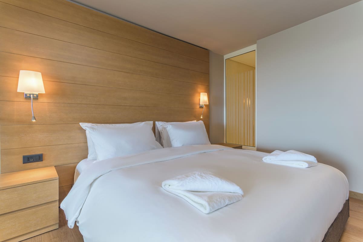 A neatly made bed with white linens and a folded towel, a wooden headboard, and two wall-mounted lamps.
