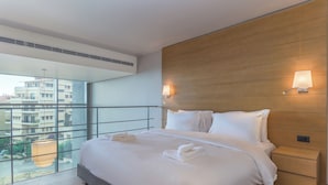 A modern hotel room with a large bed, two bedside tables, a wooden headboard, and a view of the city through floor-to-ceiling windows. - Jounieh Living - Residences by Local Host (Jounieh)