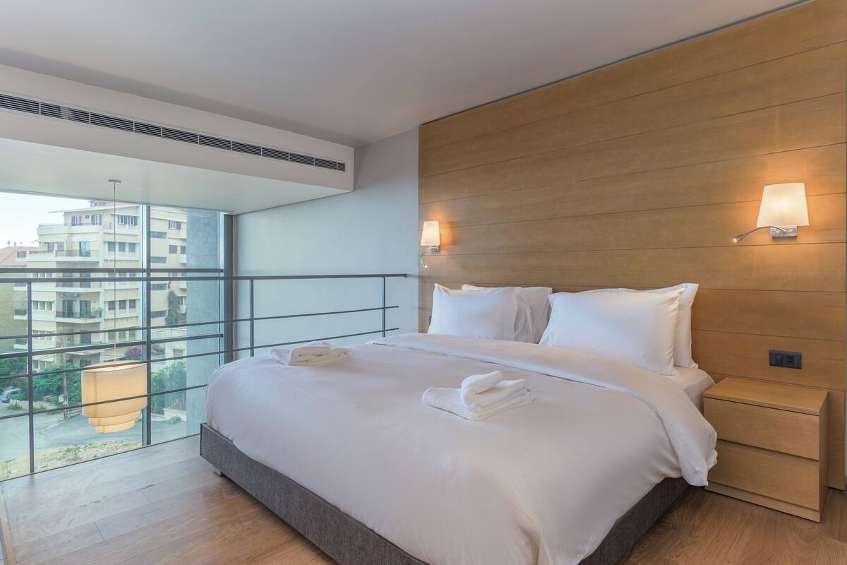 A modern hotel room with a large bed, two bedside tables, a wooden headboard, and a view of the city through floor-to-ceiling windows.