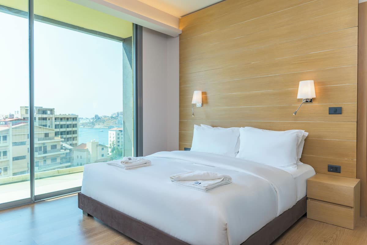 A hotel room with a large bed, two bedside lamps, a wooden headboard, and a view of the city through large windows.