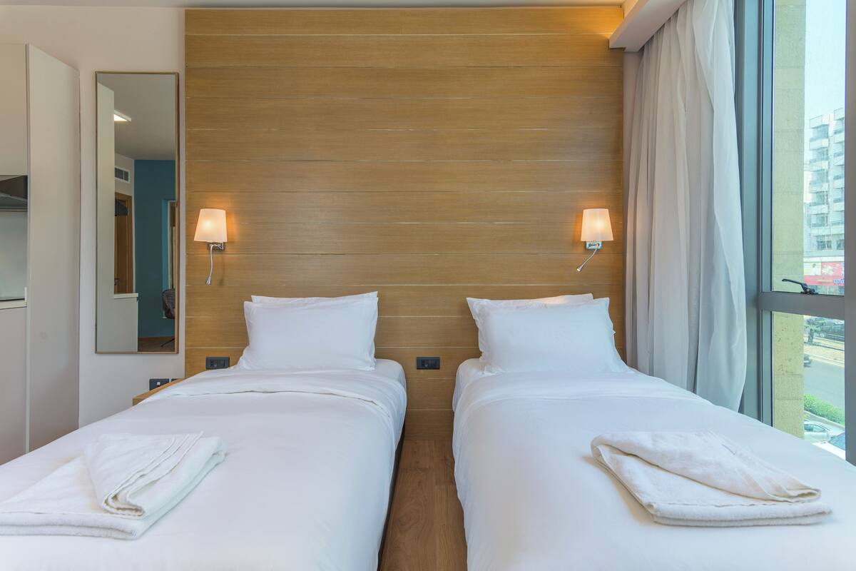 A hotel room with two beds, wooden headboard, bedside lamps, and a large window with curtains.