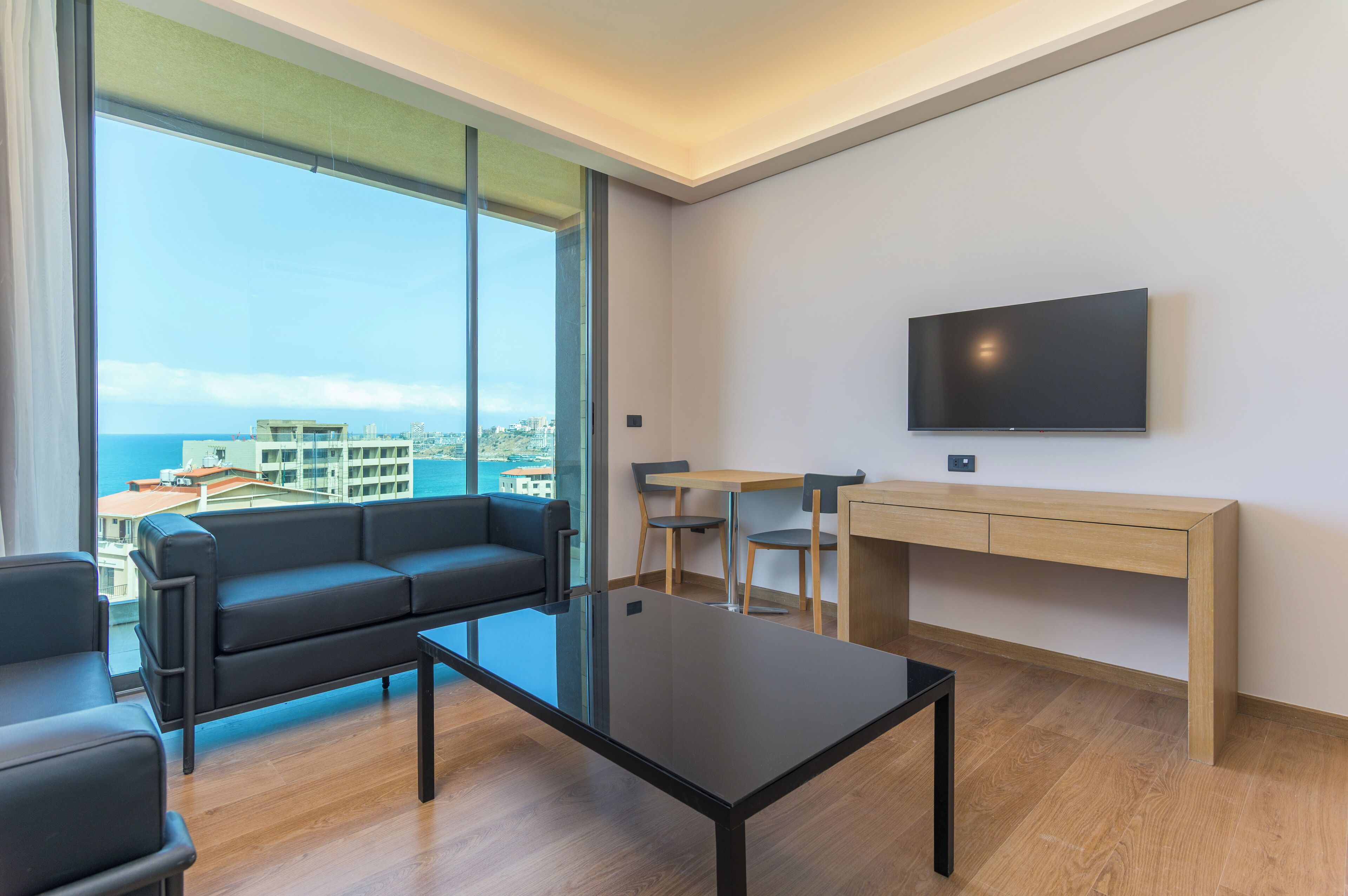 A modern living room with a black sofa, a glass coffee table, a wooden desk, and a TV mounted on the wall.