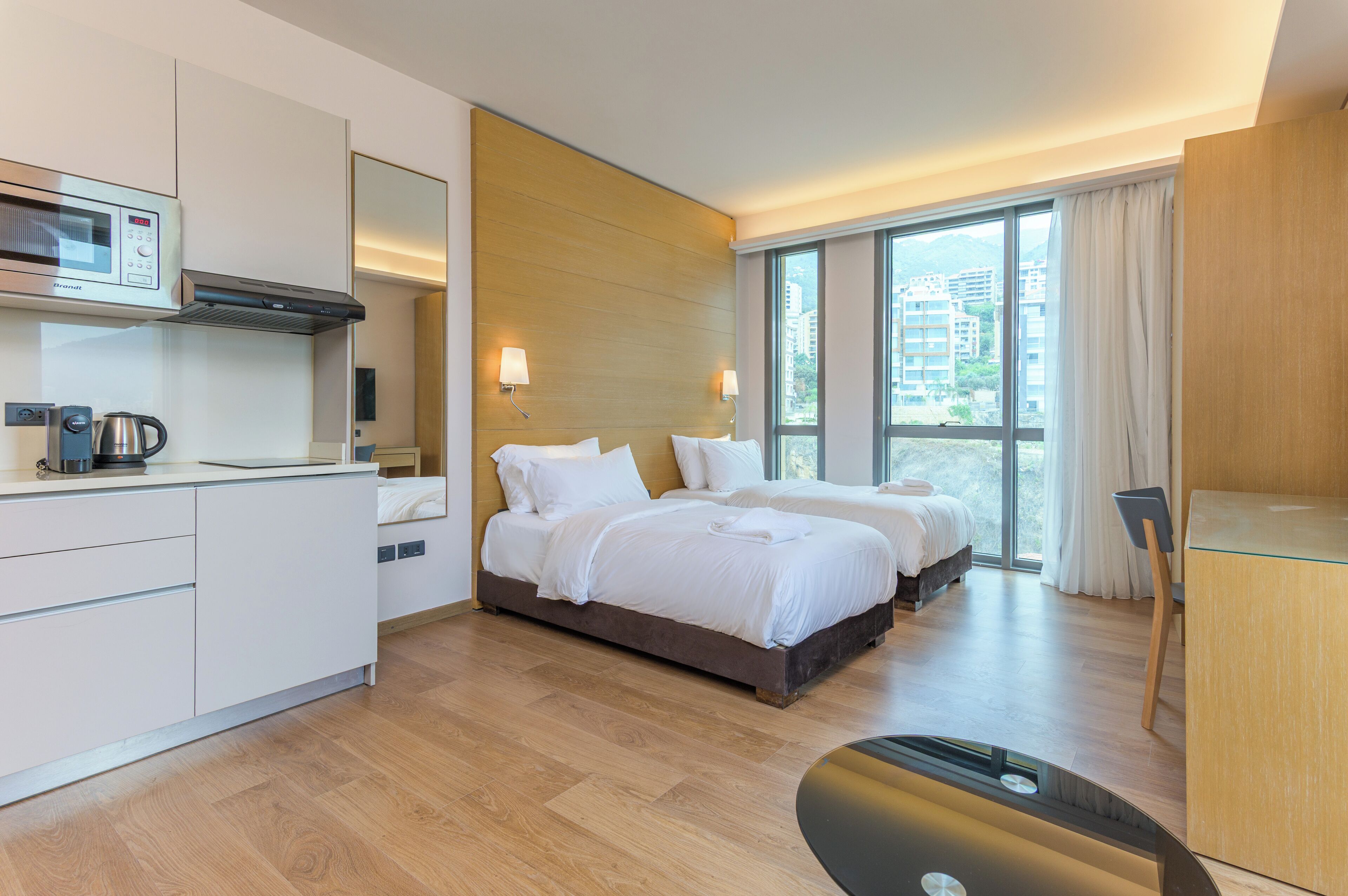 Una habitación de hotel moderna con kitchenette, dos camas, un ventanal con vistas y un escritorio.
