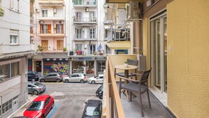 A balcony with a table and chairs, overlooking a street with parked cars and a building with balconies and air conditioning units. - Eleanor by halu! (Thessaloniki)