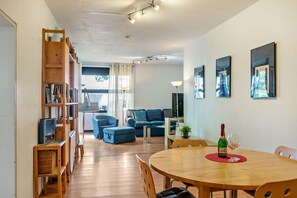 Ein Wohnzimmer mit einer Couch, einem Esstisch mit Stühlen, einem Bücherregal und einem Fenster mit Vorhängen.