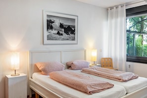 Ein Doppelbett mit Kopfteil, zwei Nachttische mit Lampen, ein gerahmtes Foto an der Wand und ein Fenster mit Vorhängen.