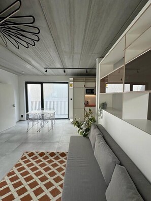 Ein modernes Wohnzimmer mit Sofa, Esstisch und Stühlen. Ein großes Fenster und eine Pflanze sind rechts zu sehen.