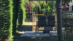 Un espace patio avec une table et des chaises sous un parasol bleu, entouré de haies bien entretenues et d’un mur de pierre.