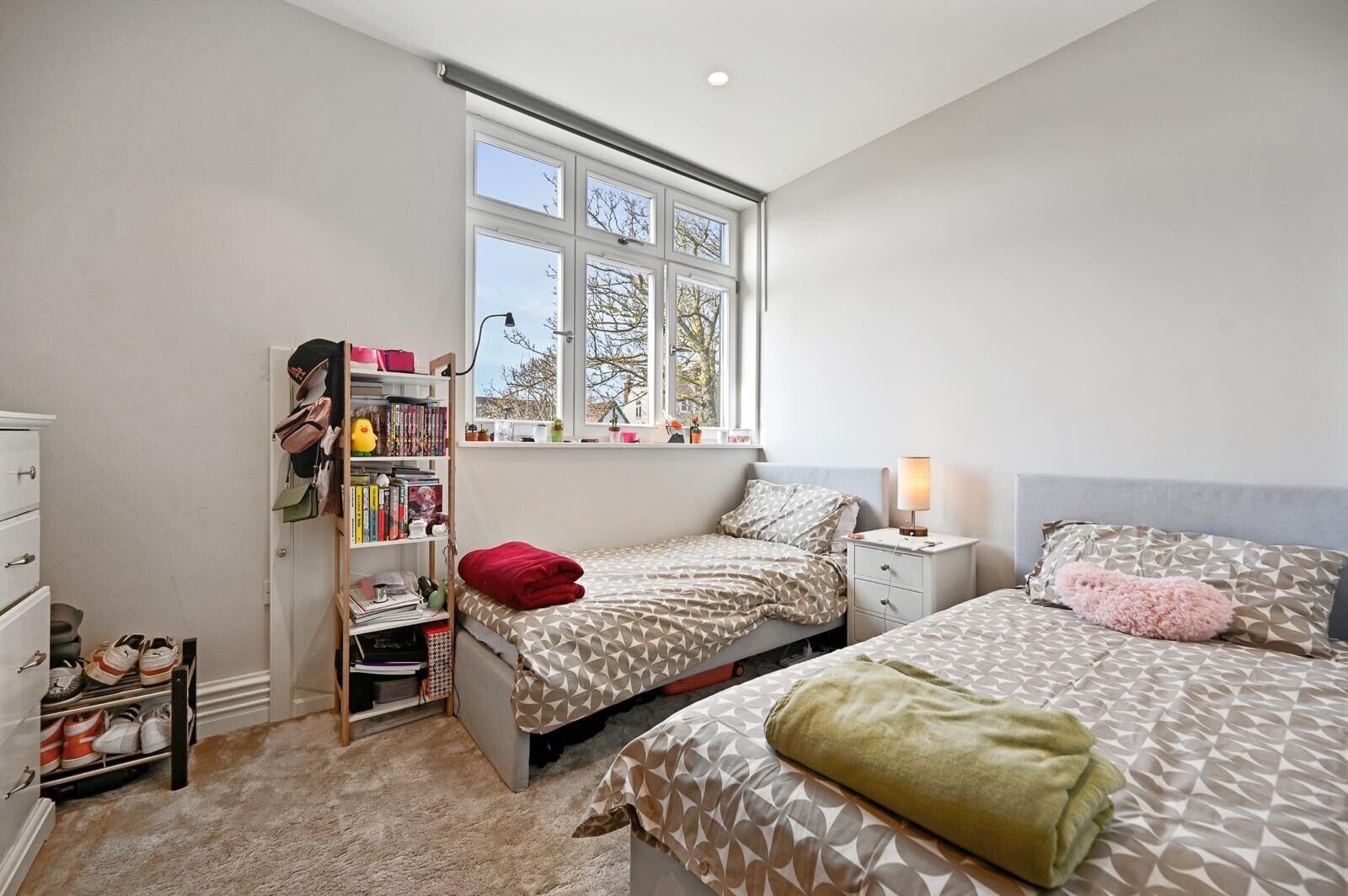 A bedroom with two beds, a bookshelf, a desk with a lamp, and a window with a view of trees.