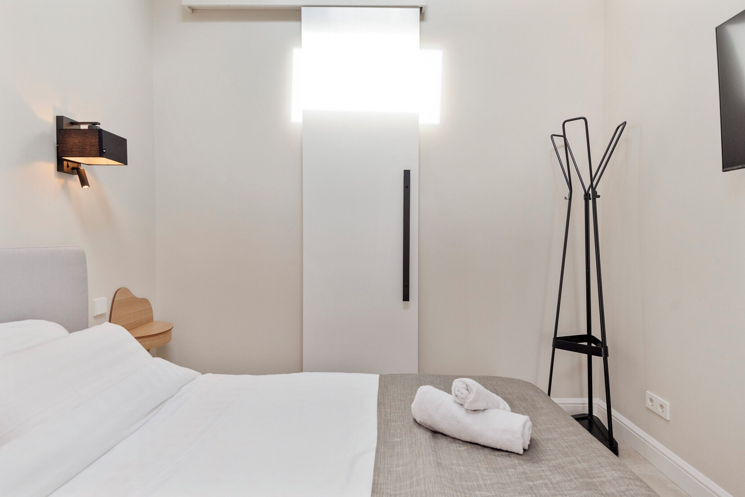 A hotel room with a bed, a nightstand, a wall-mounted light, a black floor lamp, and a folded towel on the bed.