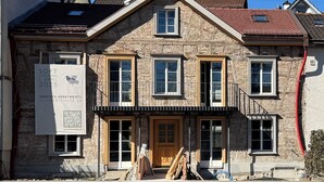 Un bâtiment en pierre avec une porte d’entrée en bois, de nombreuses fenêtres et un balcon. Une pancarte indique une ouverture en mode test prévue le 1er avril 2025.