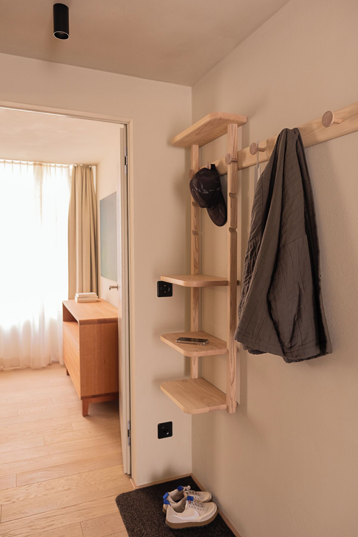 Habitación de hotel con un estante de madera, un perchero y un par de zapatillas deportivas sobre un felpudo.