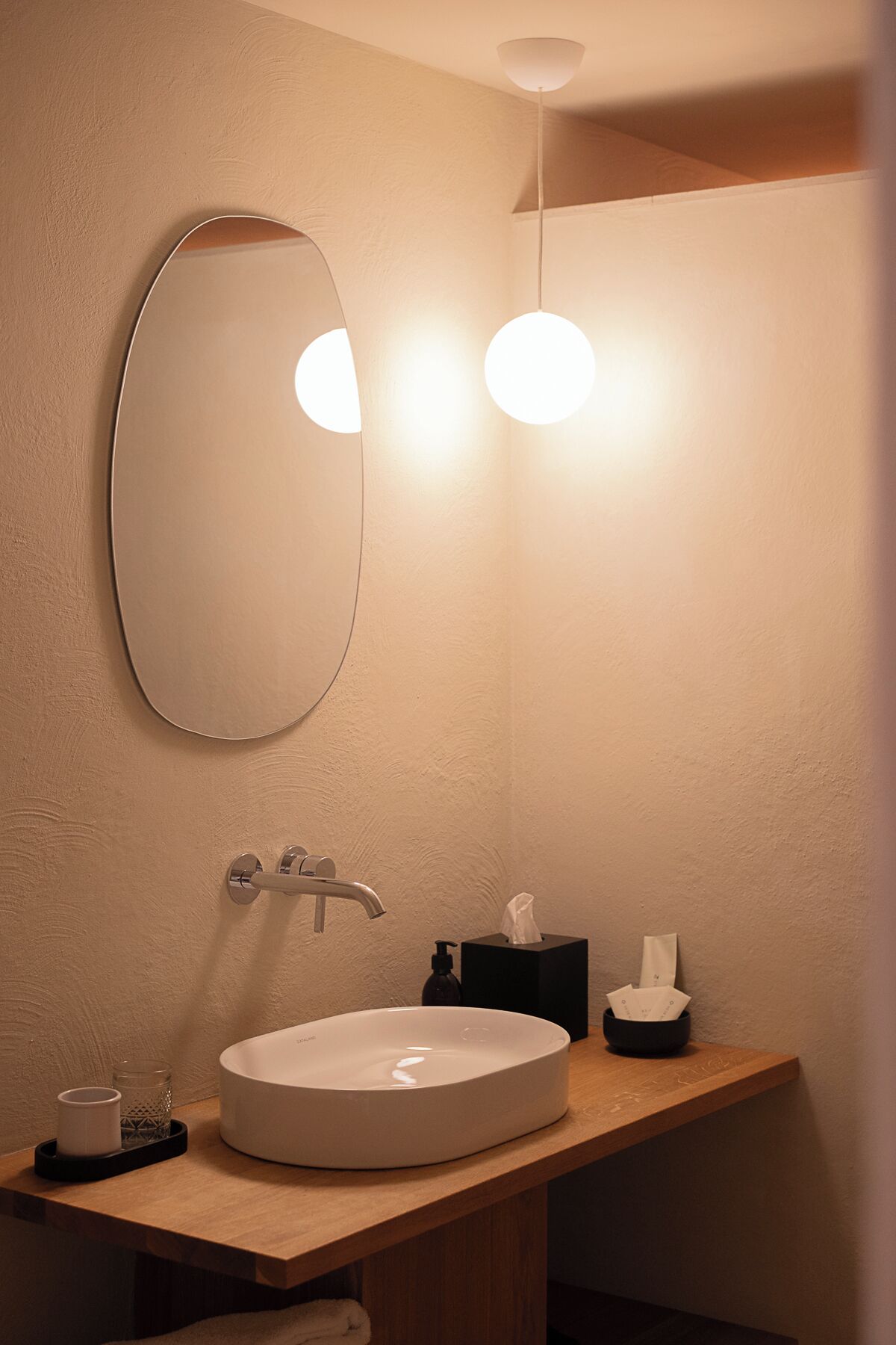 A bathroom with a round mirror, a white sink, a towel, a glass, and a black soap dispenser.