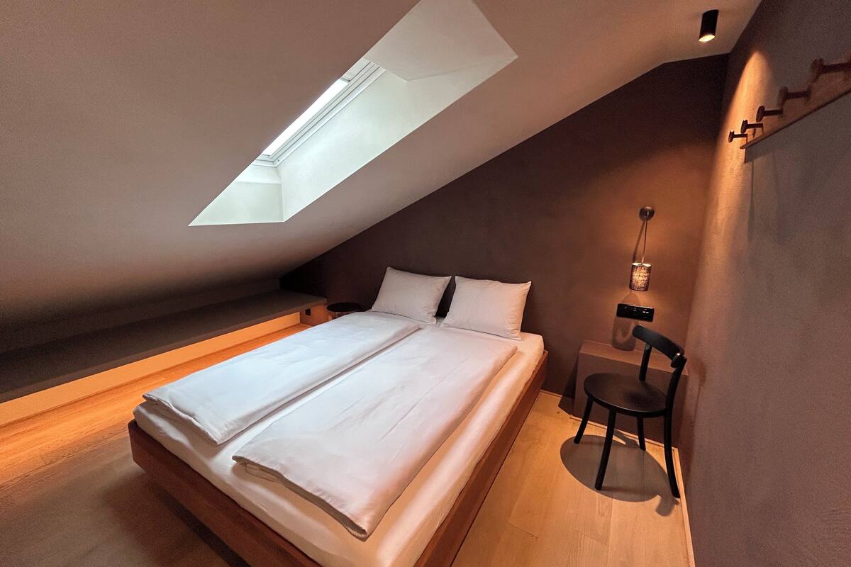 A bedroom with a skylight, a bed, a bedside table, and a chair.