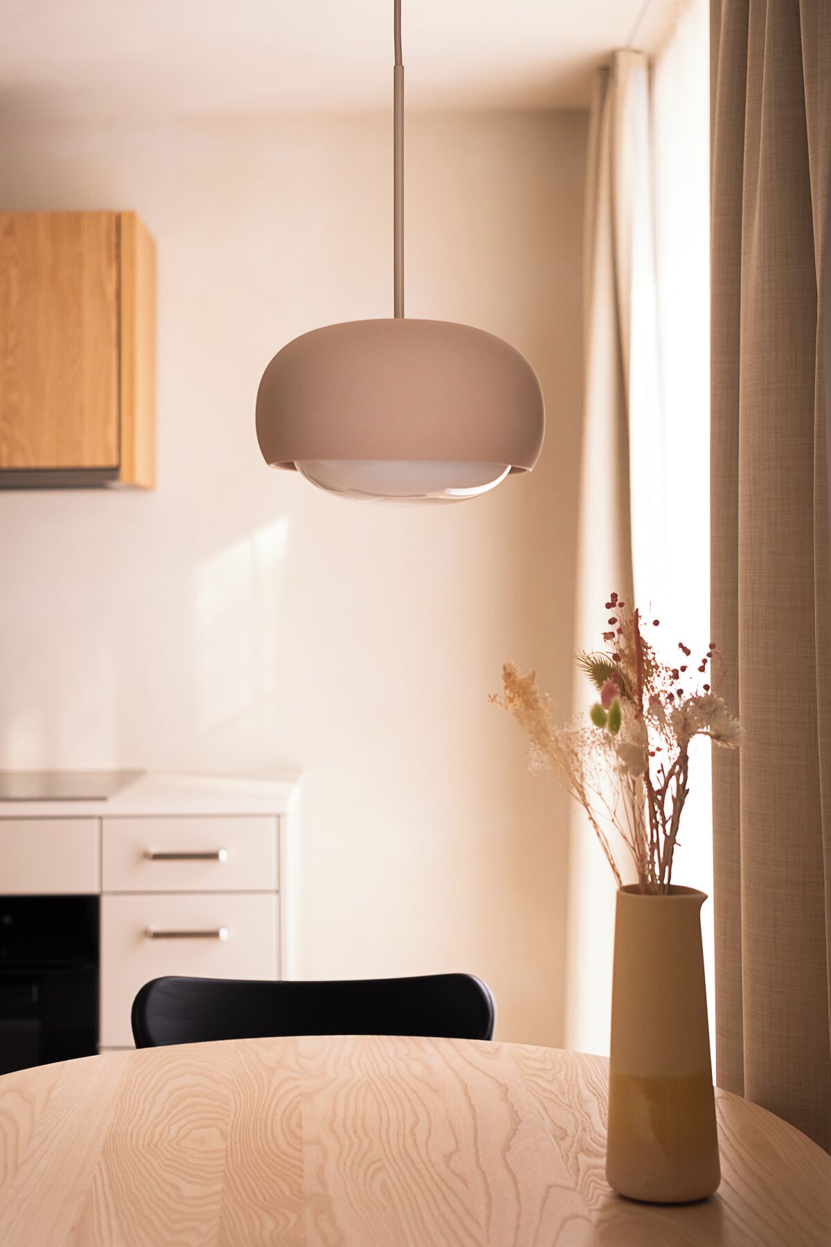 A dining area with a table, chairs, a vase with dried flowers, and a hanging pendant light.