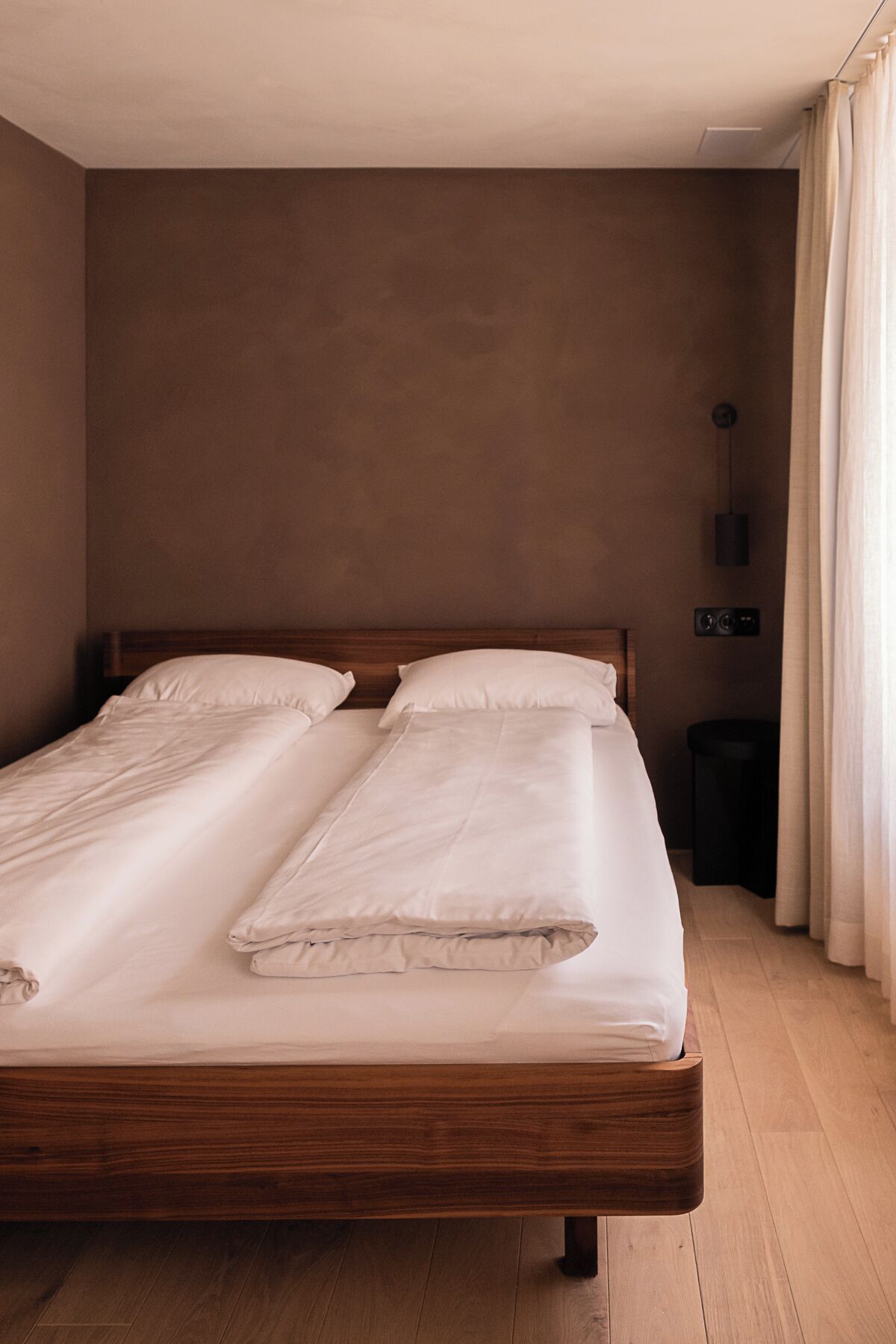 A double bed with a wooden frame and white bedding in a room with a brown wall and wooden flooring.