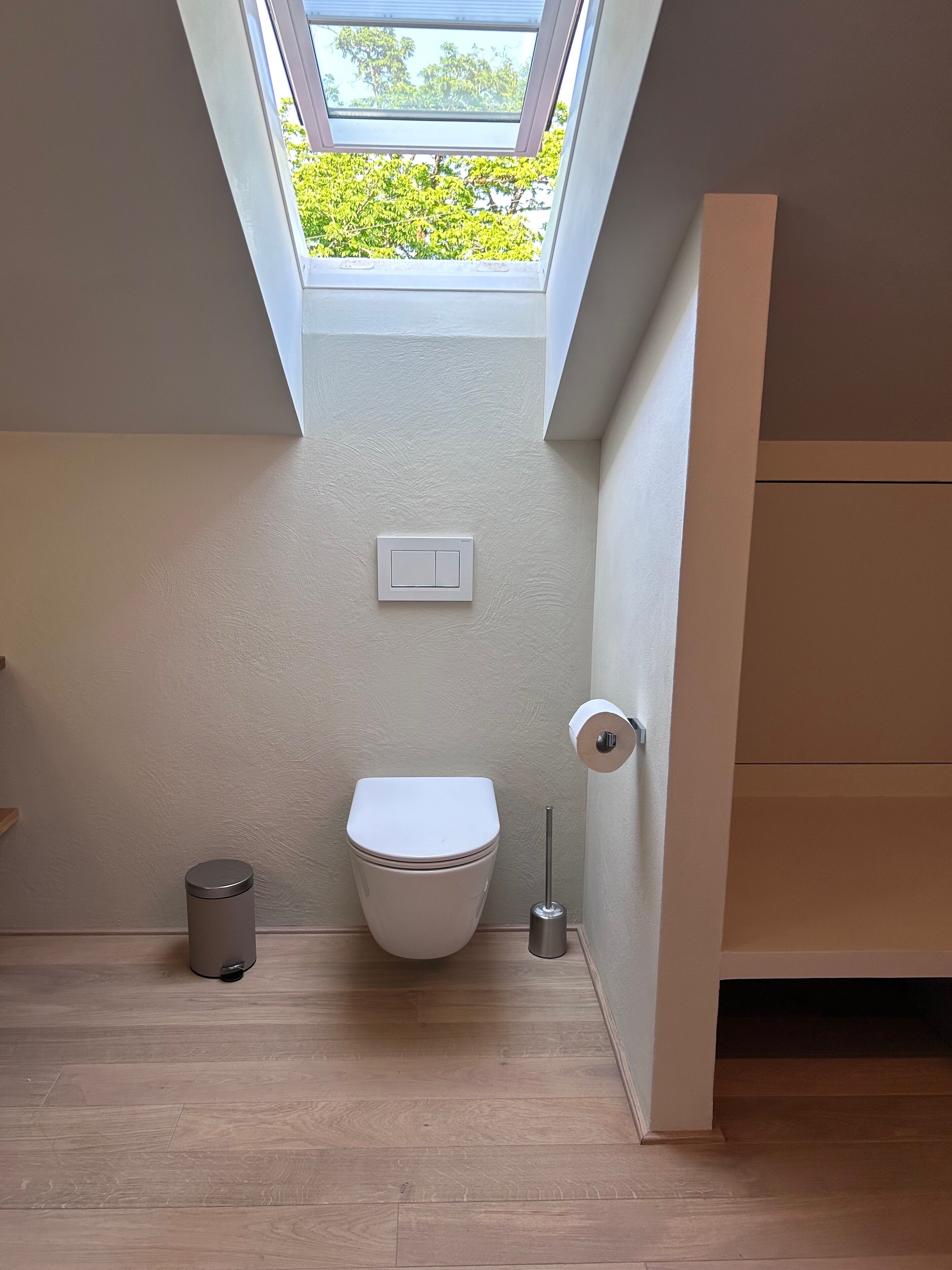 A compact bathroom with a skylight, a toilet, a trash bin, and a toilet paper holder.