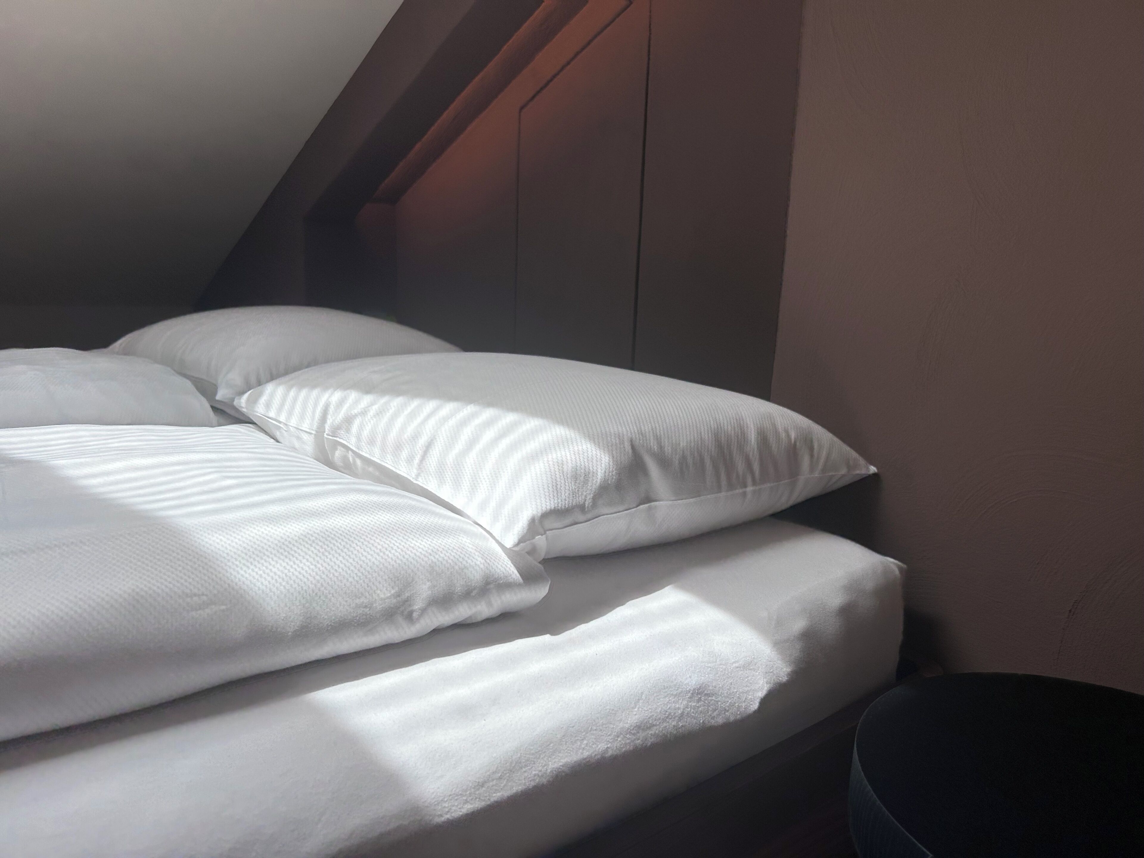 Een net opgemaakt bed met witte lakens in een kamer met schuine plafonds en een donkere wand.