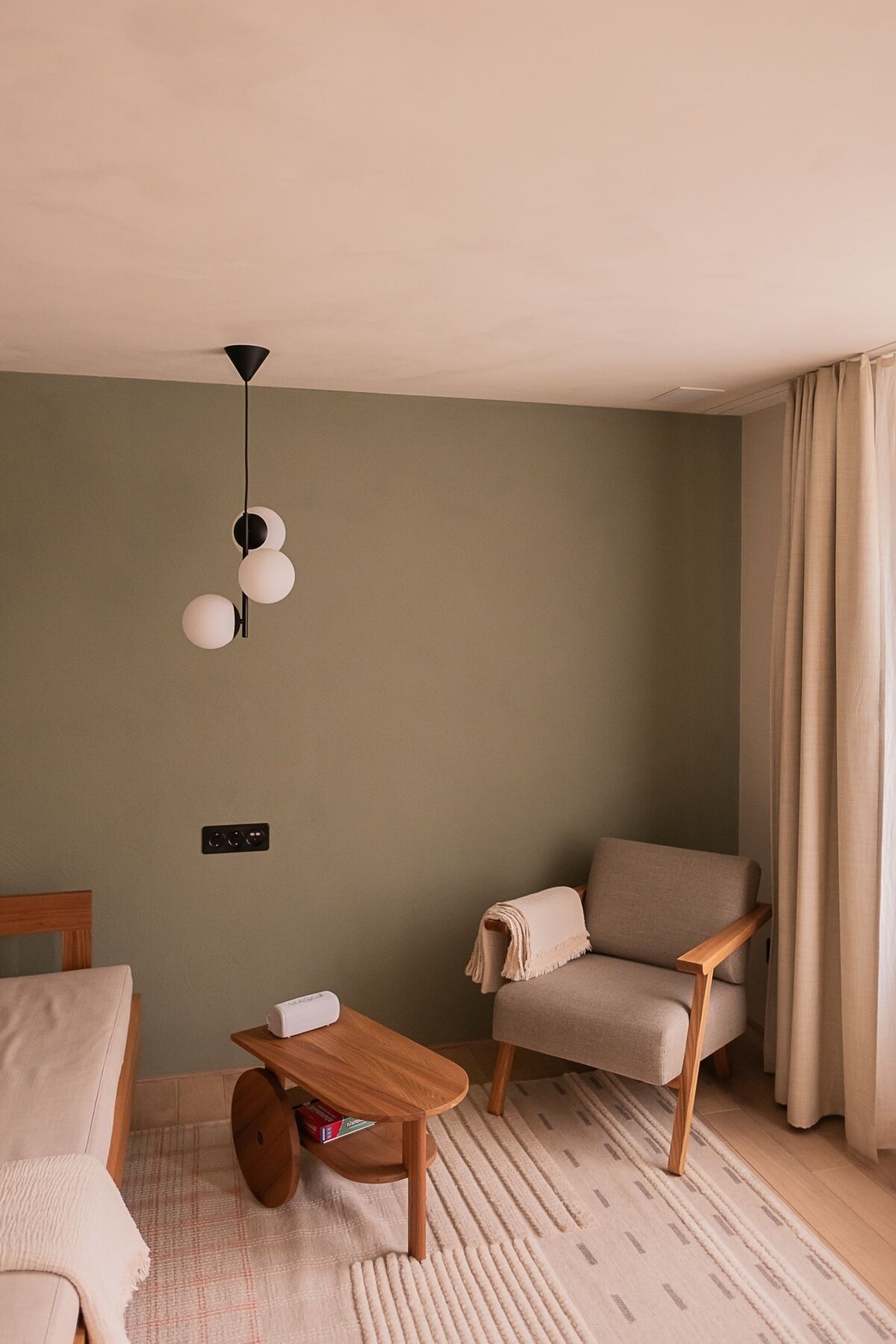 Ein gemütliches Zimmer mit einem Bett, einem Holztisch, einem beigen Sessel, einer Pendelleuchte und einer Steckdose an der Wand.