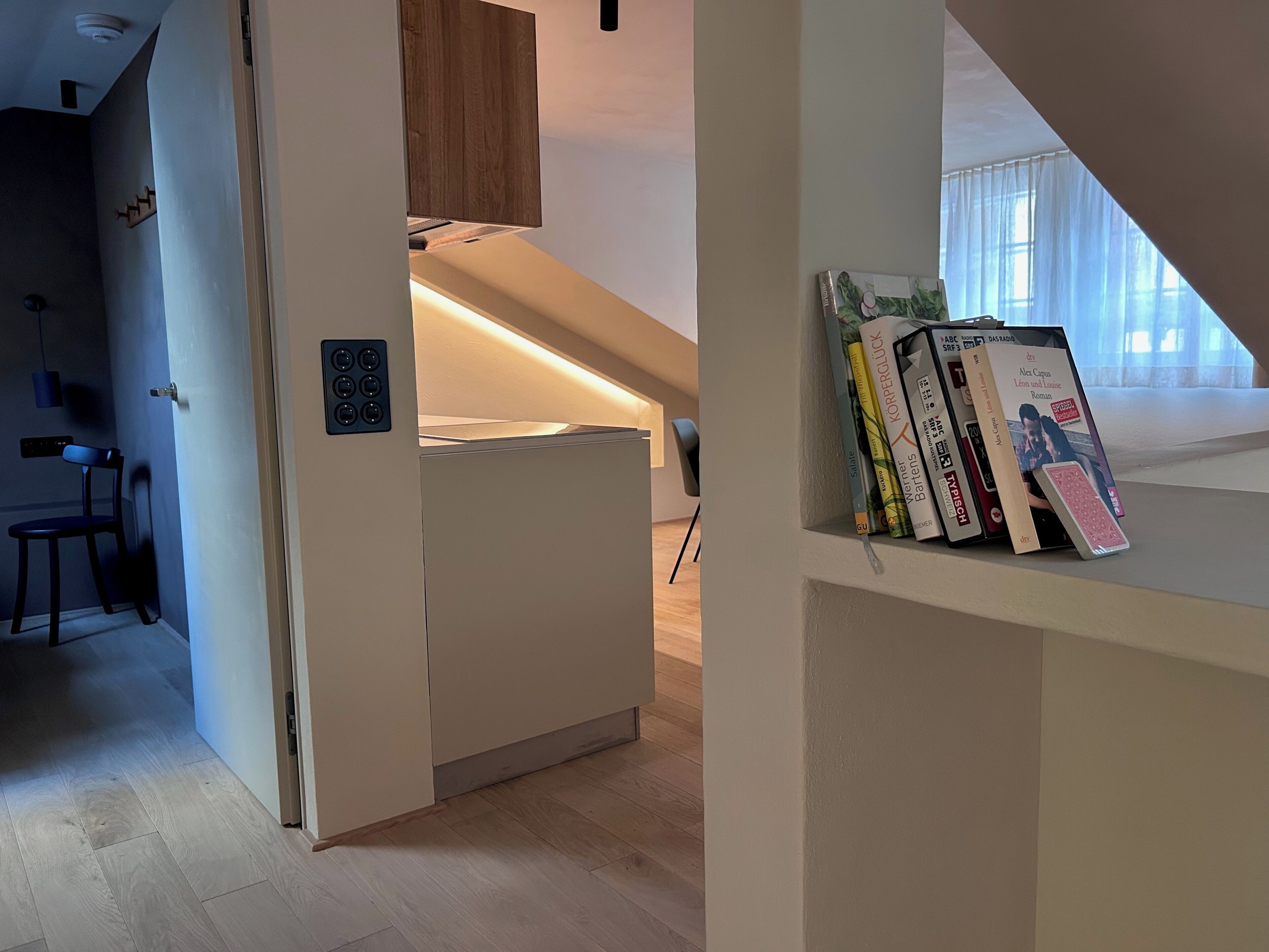 A modern, compact living space with a kitchenette, a bookshelf with books, and a view of a room with a window.