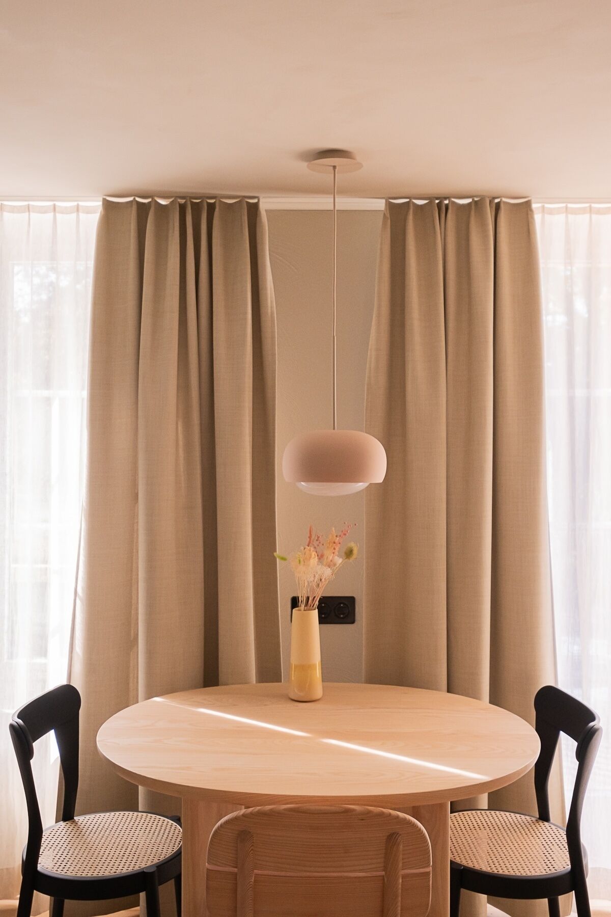 Een ronde houten eettafel met twee zwarte stoelen, een vaas met bloemen en een hanglamp.