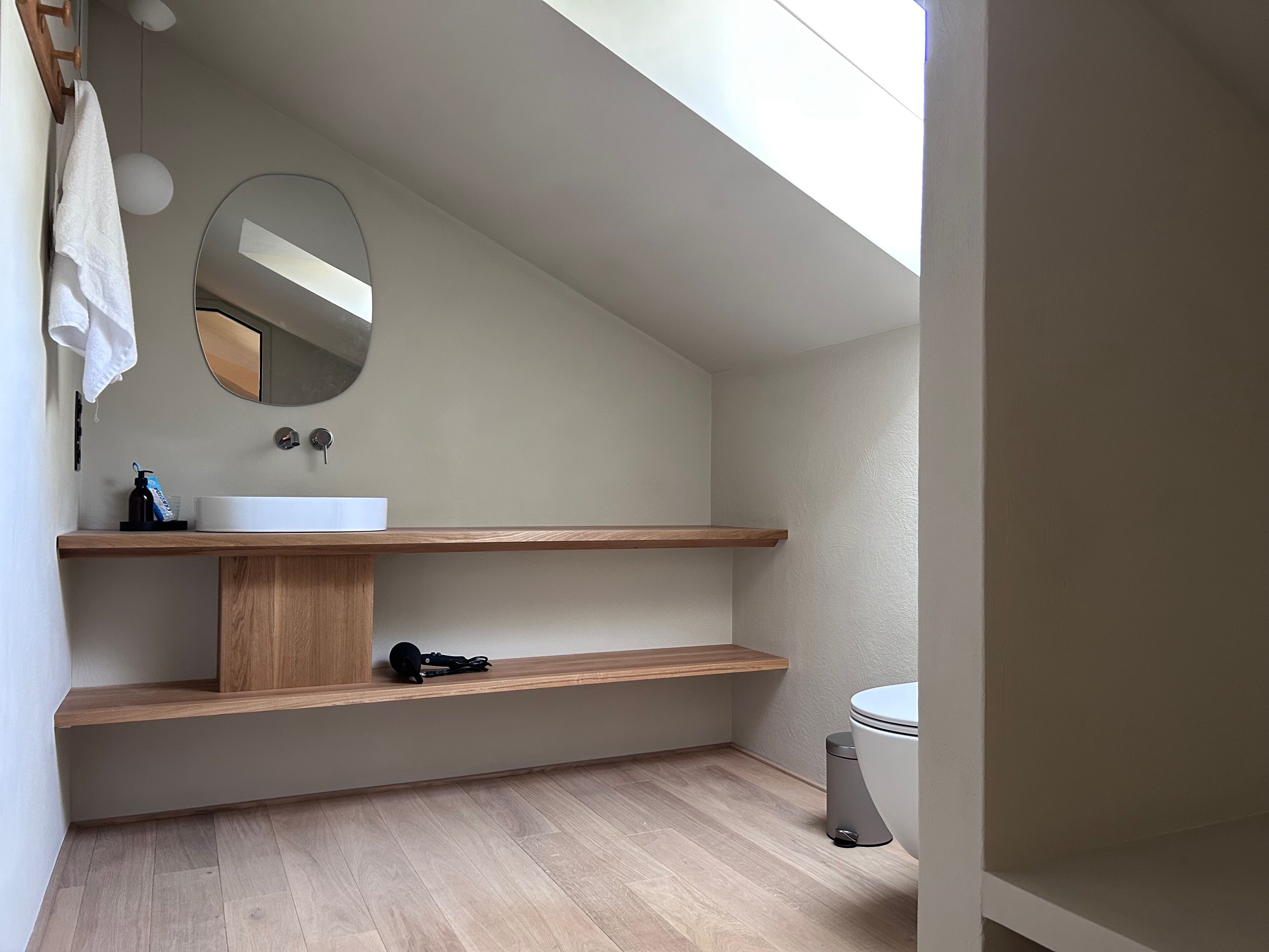 A modern bathroom with a white sink, wooden shelves, a mirror, and a toilet.