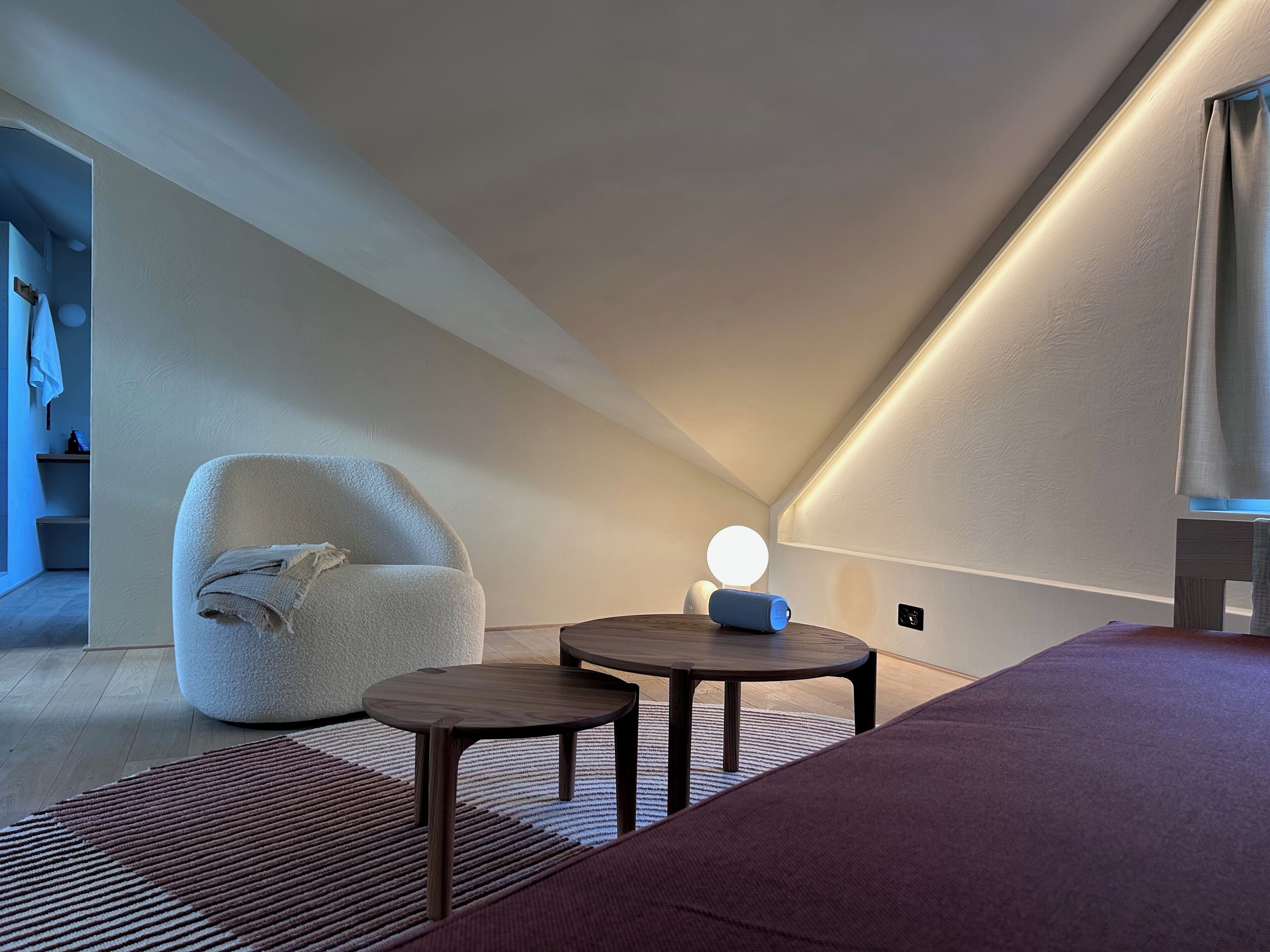 Un dormitorio moderno y luminoso en el ático con una silla acogedora, una mesita redonda y una cama.
